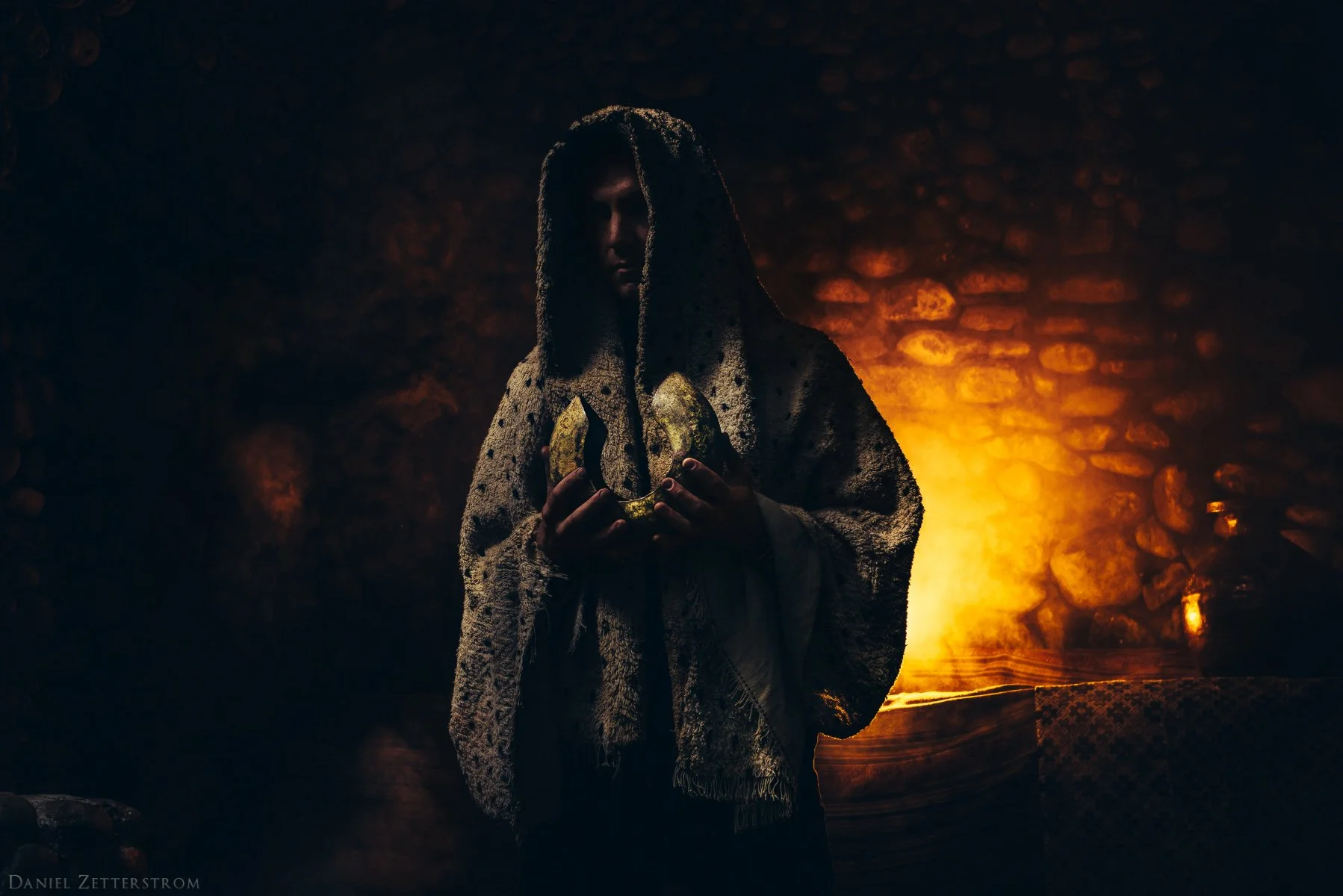 A person with long dreadlocks draped over their face, wearing a textured cloak, holding a golden object in both hands, standing in dim lighting with a warm glow behind them. Random Rab