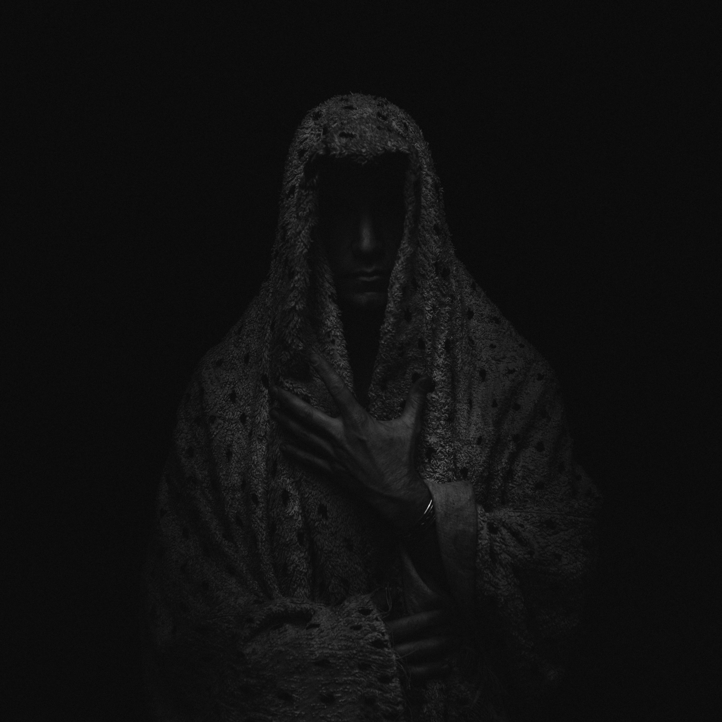 A dark, moody black and white portrait of a person with a hooded garment, their face partially obscured in shadow, holding their hand over their chest. Random Rab