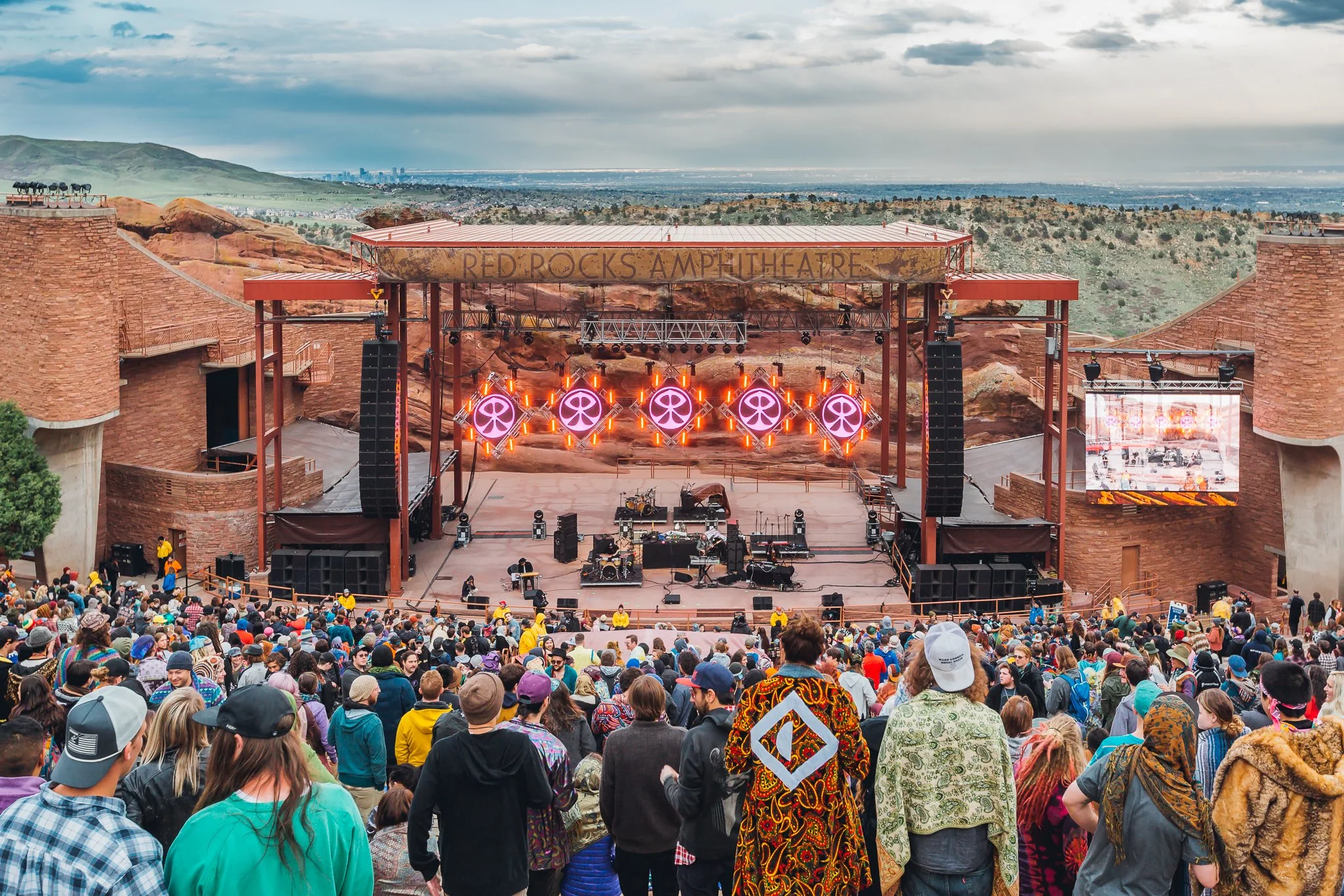 View of Red Rocks Amphitheatre with a large crowd in front, stage set with purple lighting and a band setup, surrounded by red rock formations and a cloudy sky.
