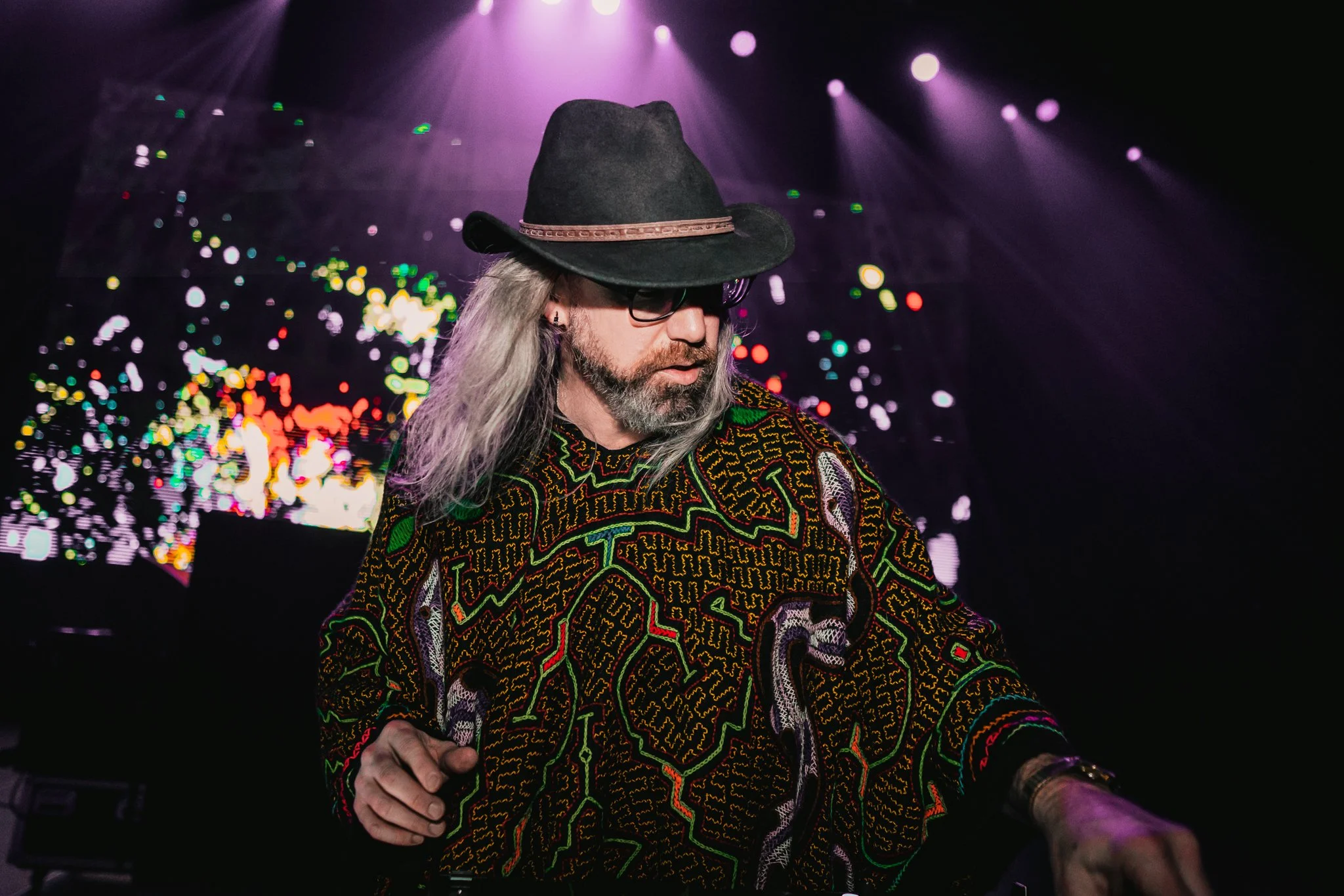 A man with long gray hair, glasses, and a beard wearing a black cowboy hat and a colorful patterned shirt, performing at a concert with purple stage lights and colorful digital backdrop. Random Rab