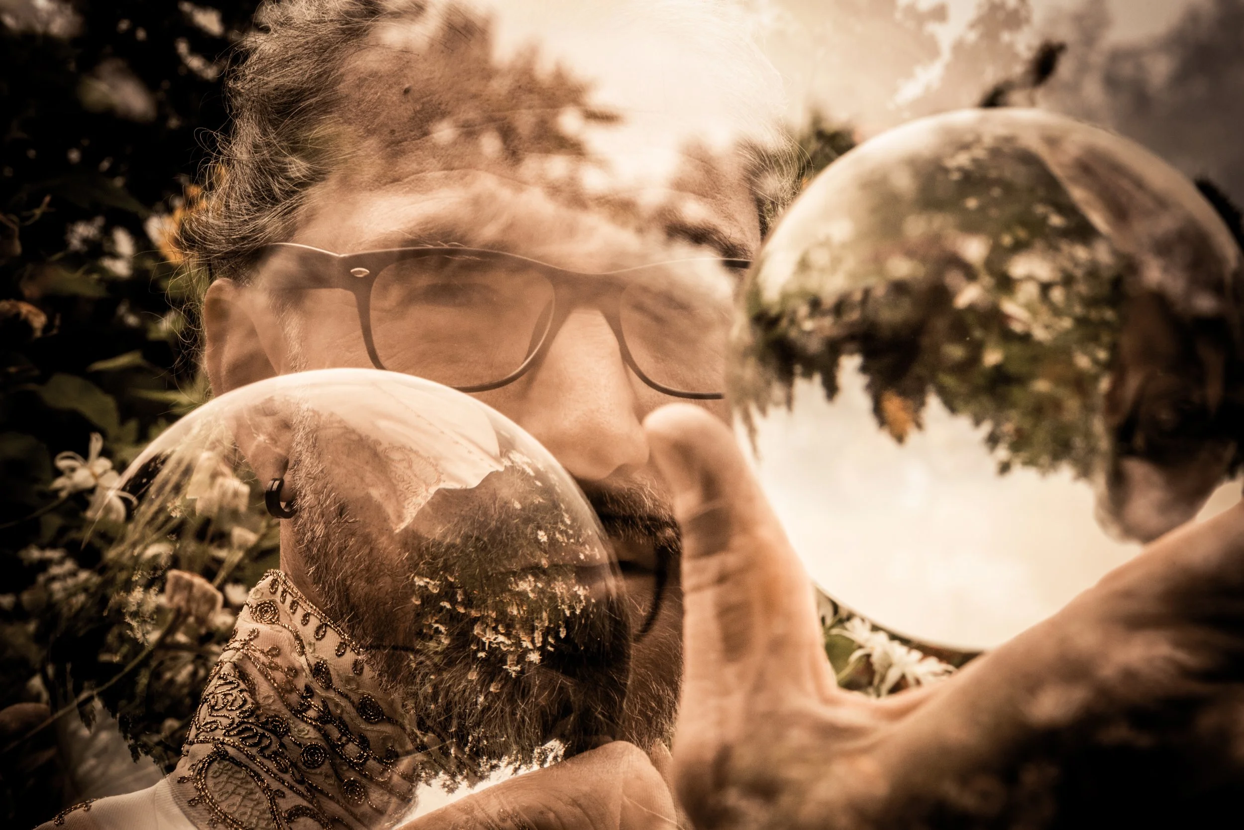 A person with glasses and a beard is looking through a transparent sphere, with trees and sky reflected on its surface. The scene appears to be outdoors during the day.