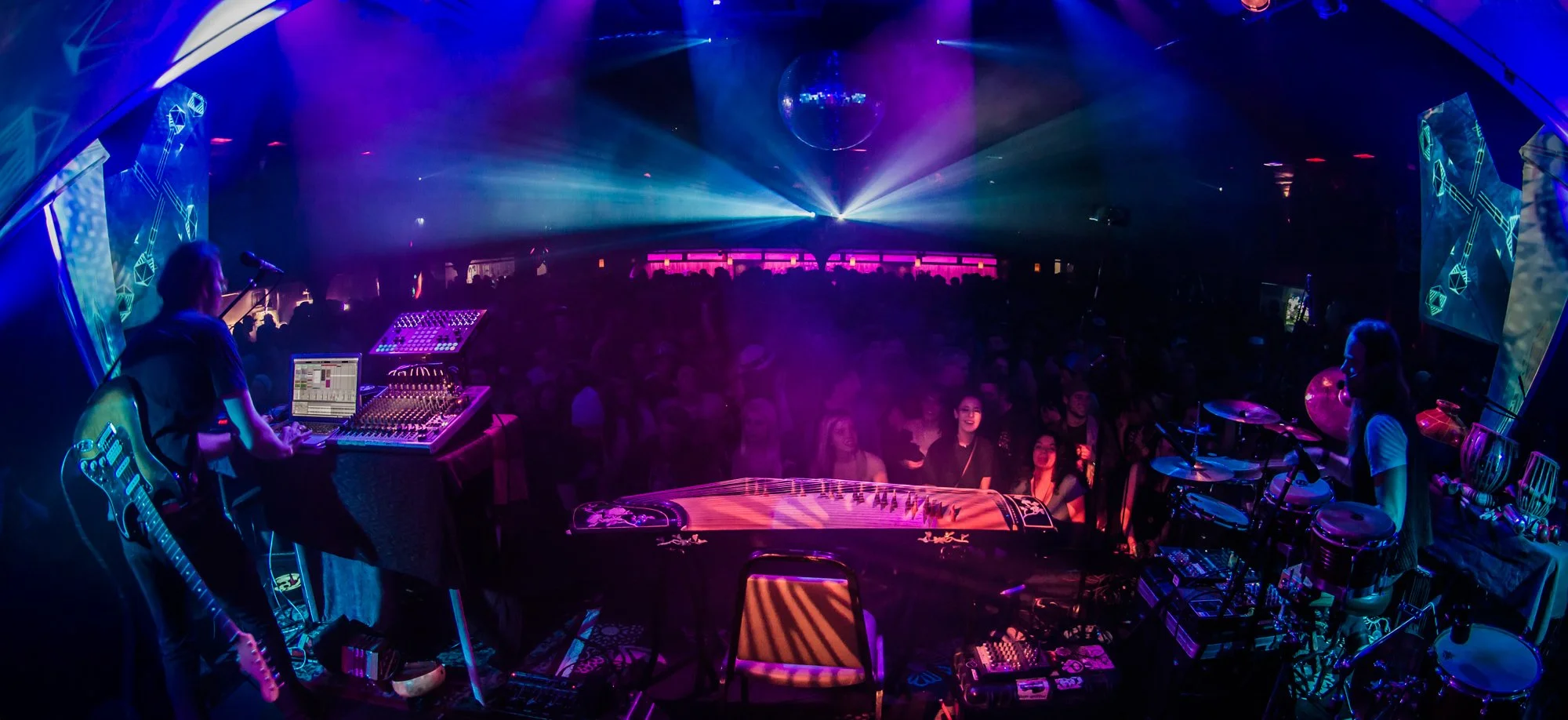 A DJ playing electronic music at a nightclub with colorful lights, a large crowd dancing, and a stage with electronic instruments and music equipment. Random Rab