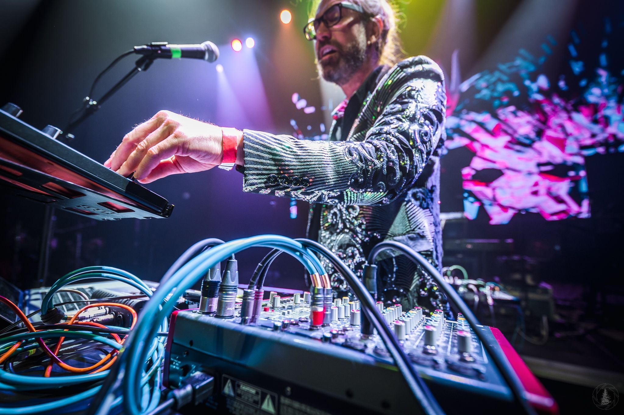 A DJ with long hair, glasses, and a beard, wearing a shiny, patterned jacket, is performing on stage. He is standing behind a turntable and mixer with colorful cables. The background features vibrant, multicolored lights and a digital screen displaying abstract visuals.