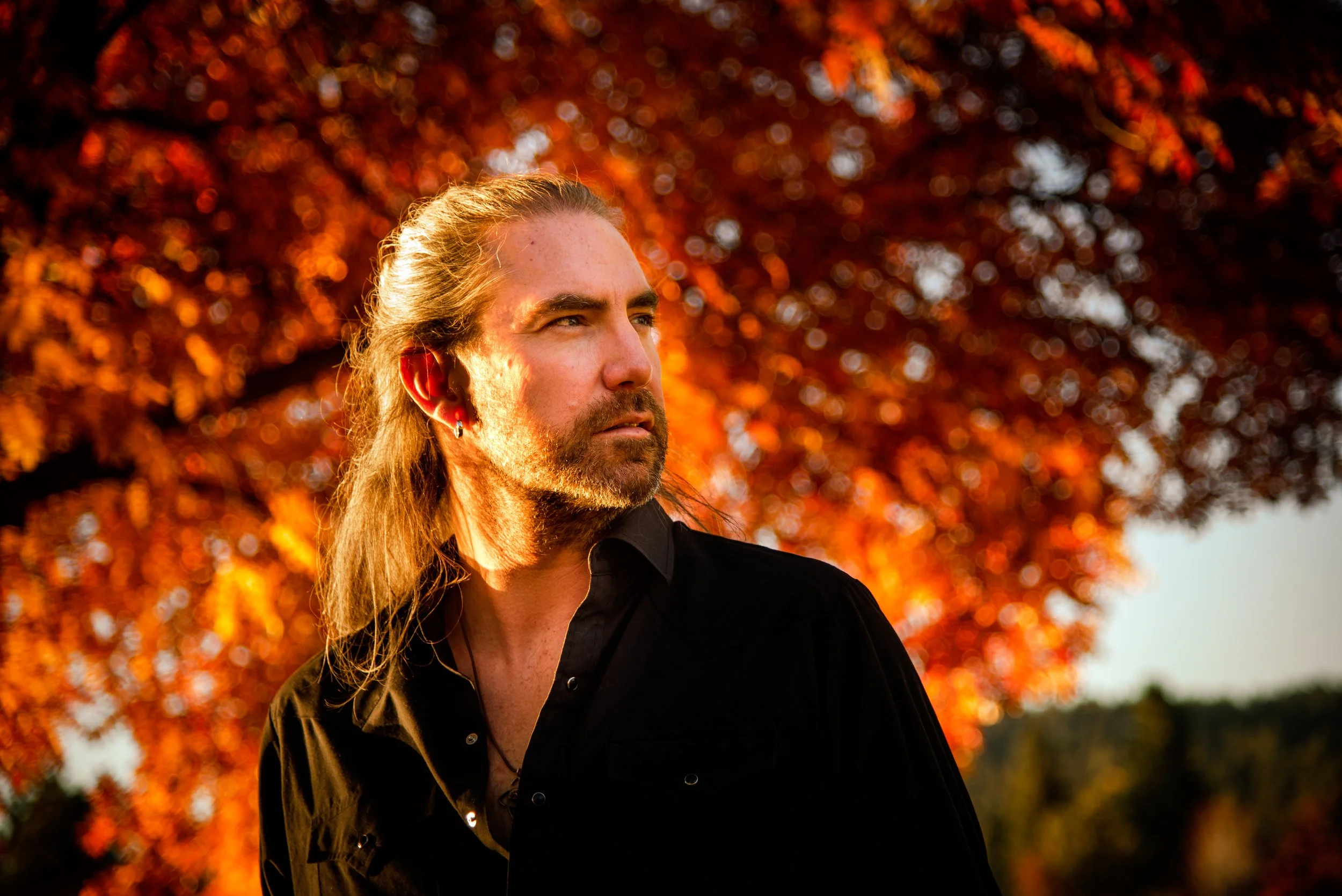 A man with long hair and a beard stands outdoors during sunset, with autumn trees with orange leaves in the background.