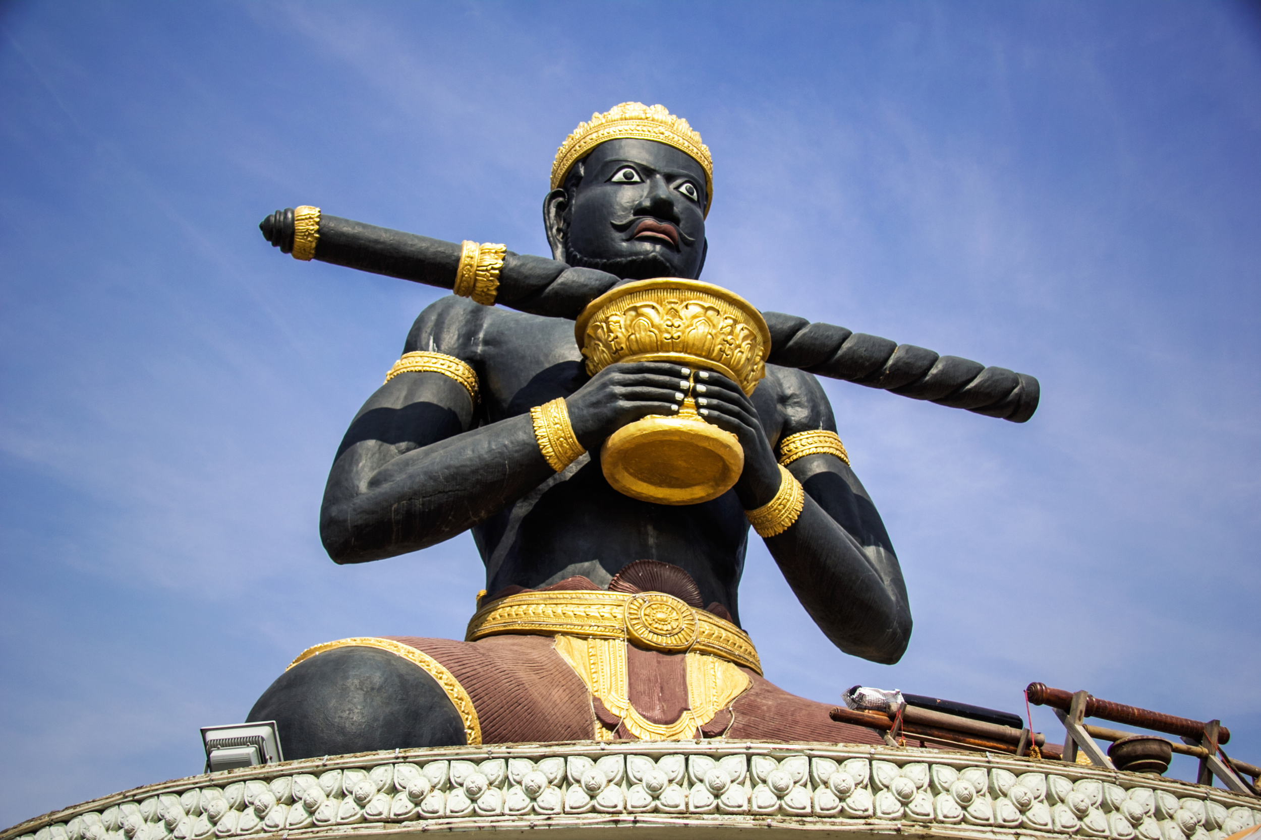 Close-up of a large black statue of a Sikh figure with a gold crown, holding a golden cup, against a blue sky.