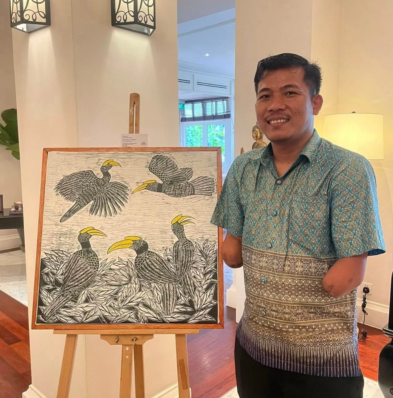 A man standing indoors next to an artwork of six hornbills, with some flying and some perched among leaves, all rendered in black and white with yellow beaks. The man is smiling and wearing a patterned turquoise shirt.