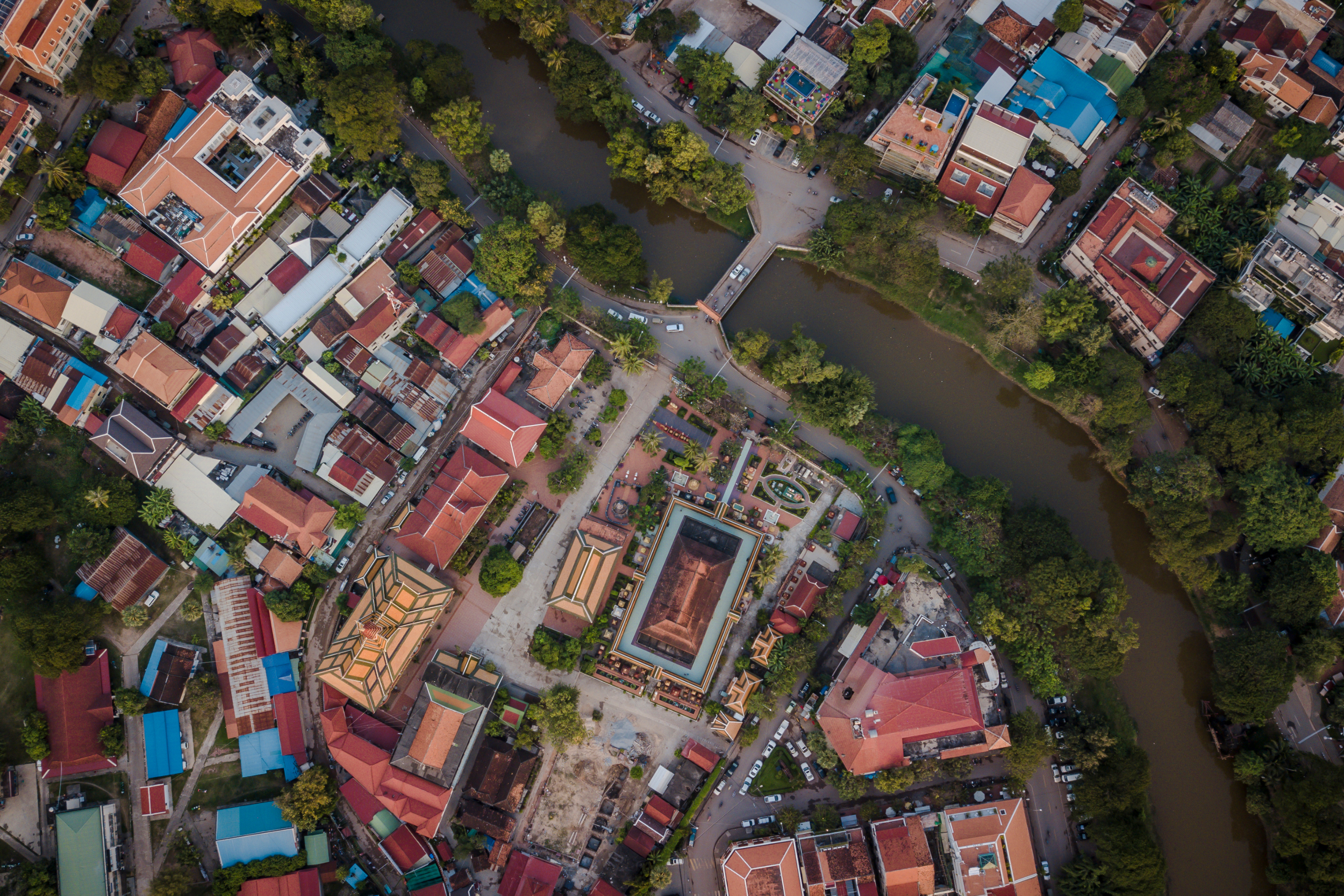 Aerial view of a city with a river running through the middle, surrounded by buildings, trees, and roads.