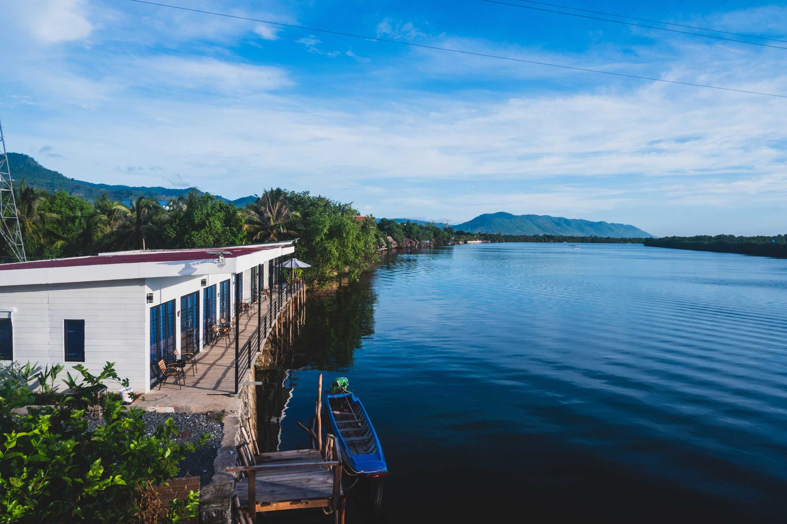 A riverside scene with a building and outdoor seating, small boat docked along the shore, lush green trees, distant mountains, and a blue sky with scattered clouds.