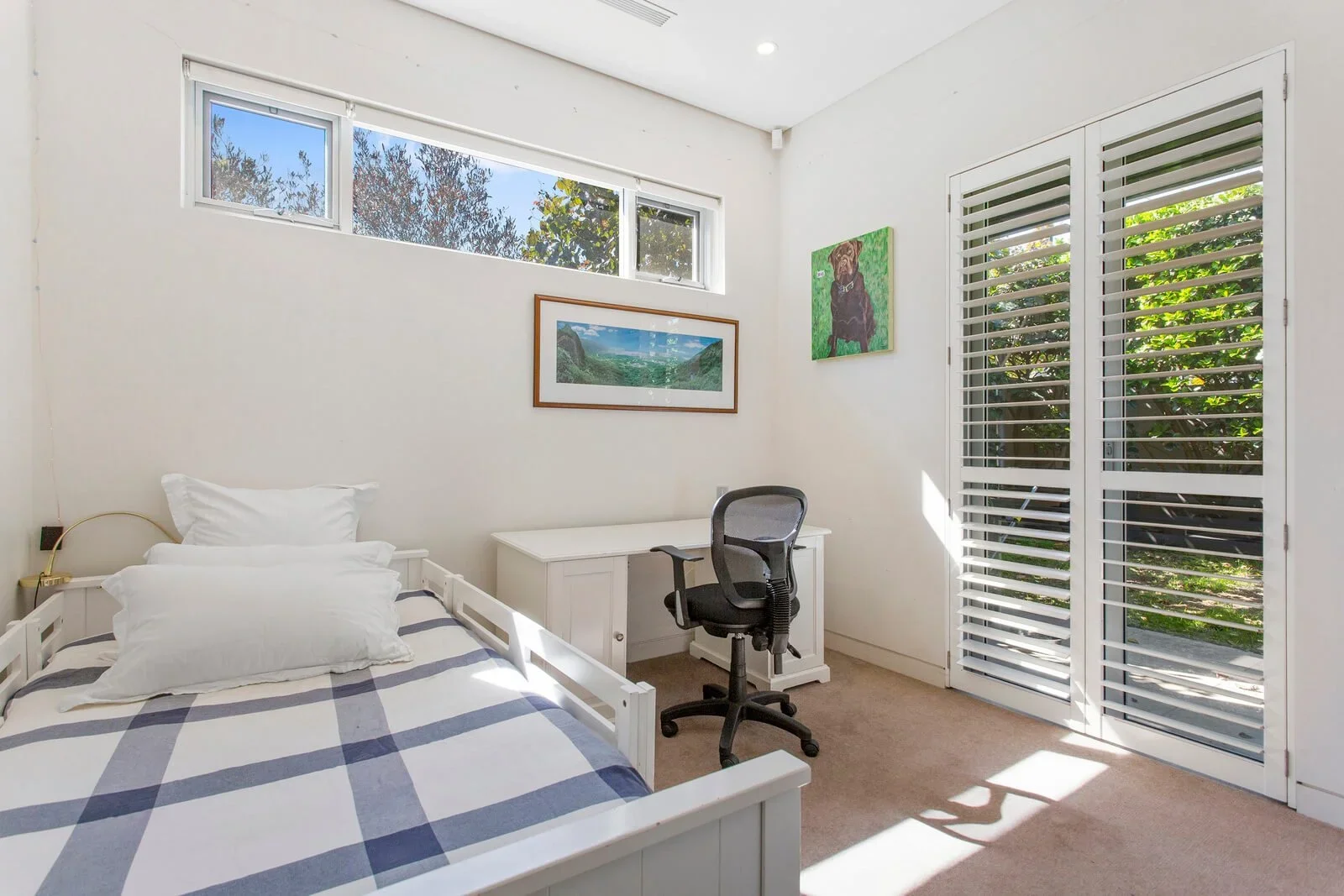 A minimalist bedroom with a white bed, black office chair, white desk, three horizontal windows, and white louvered doors. Artwork hangs on the wall, and sunlight streams through the doors.