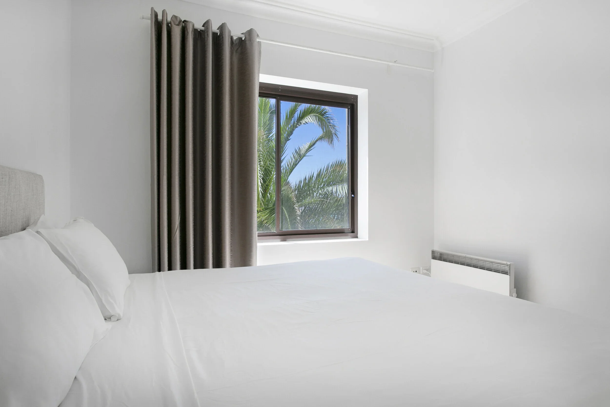 A bedroom with a neatly made white bed, a window with a view of palm trees, and brown curtains.