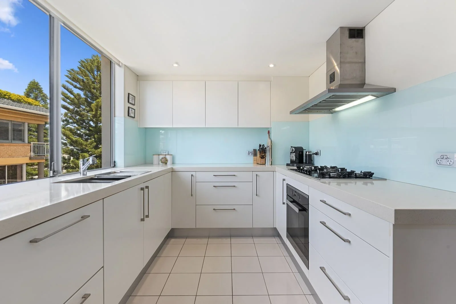 Modern kitchen with white cabinets, beige tile floor, and large window showing blue sky and trees outside. Contains a stainless steel stove, oven, coffee machine, and various kitchen utensils.