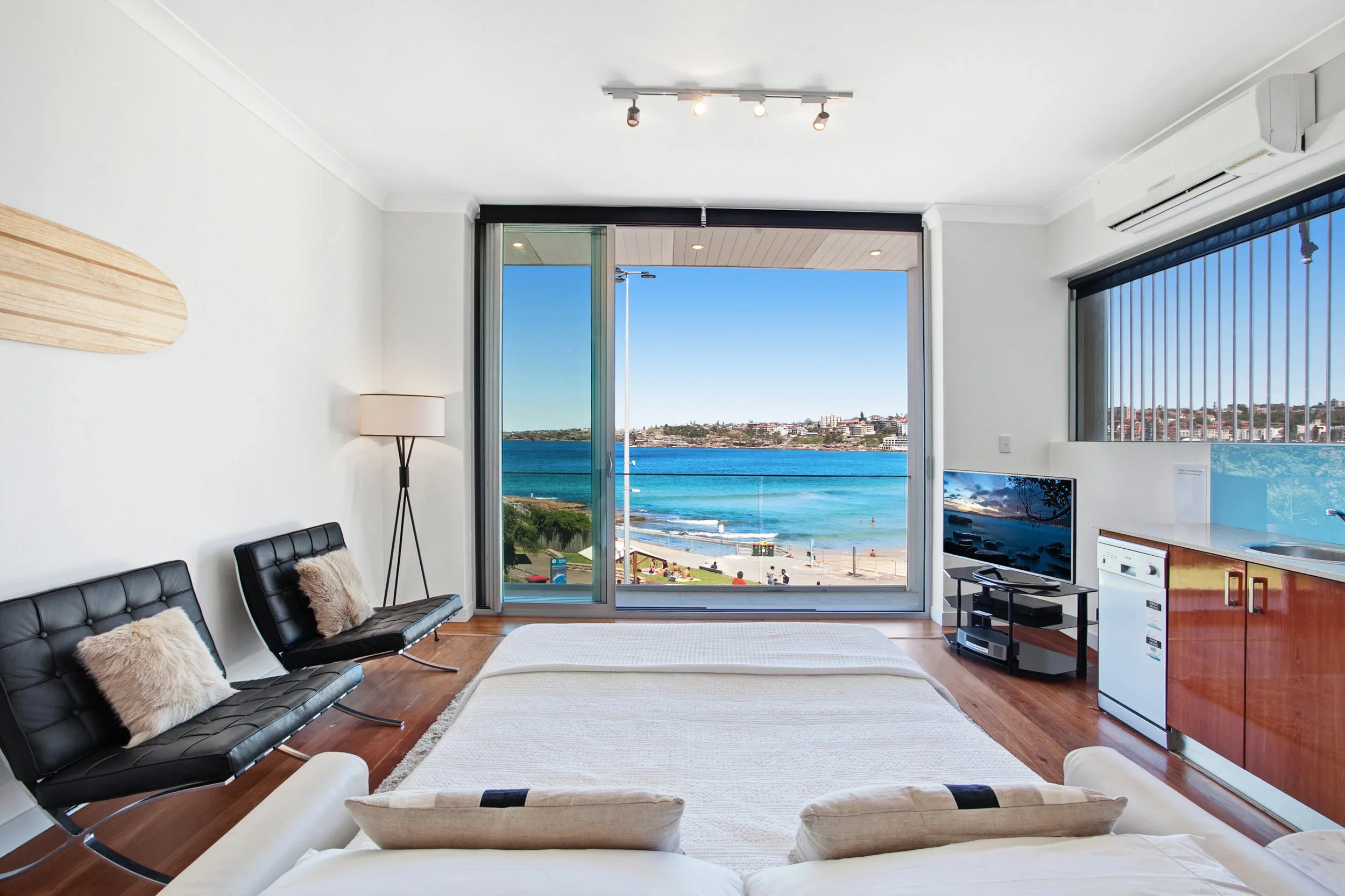 Living room with large sliding glass door opening to a balcony with ocean view, two black chairs with fur pillows, a white sofa, a flat-screen TV, and a kitchen area with a window and blinds.