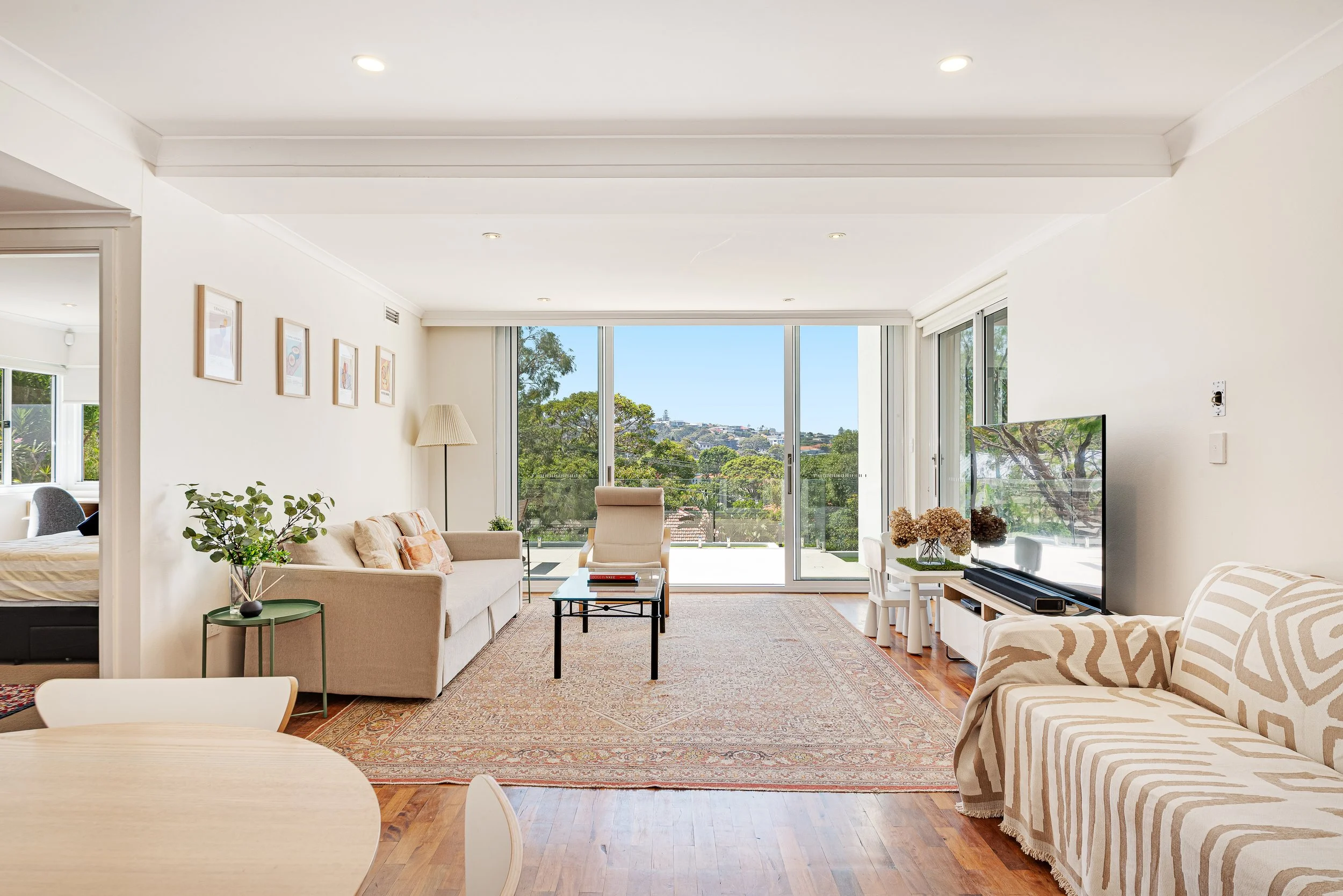 Living room with a large white sofa, black armchair, glass coffee table, and a wooden outdoor balcony with trees outside.