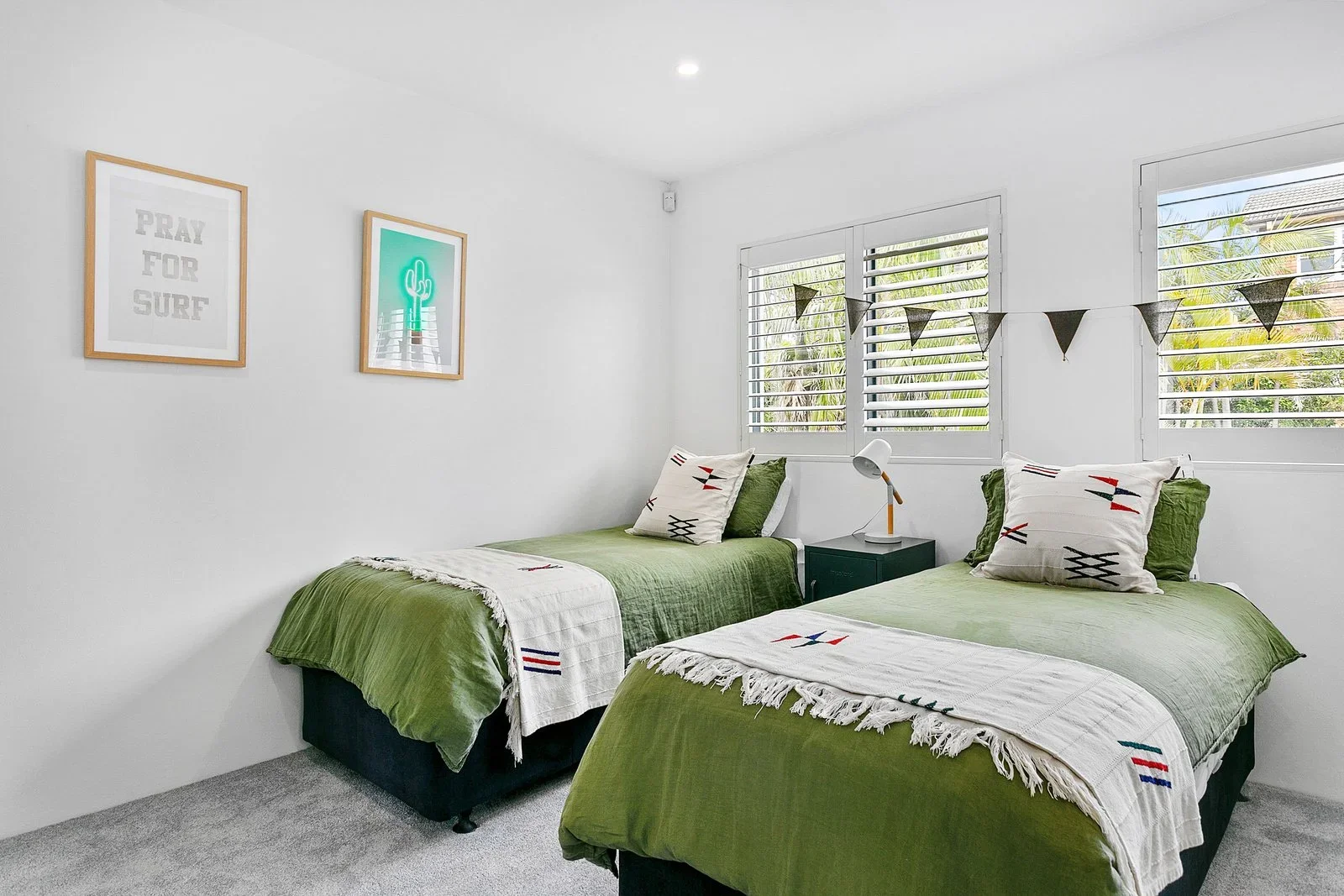A bedroom with two twin beds covered in green bedding and decorated with pillows and white throws with colorful patterns. The room has white walls, a gray carpet, three windows with white shutters, and wall art with quotes and drawings. A small teal