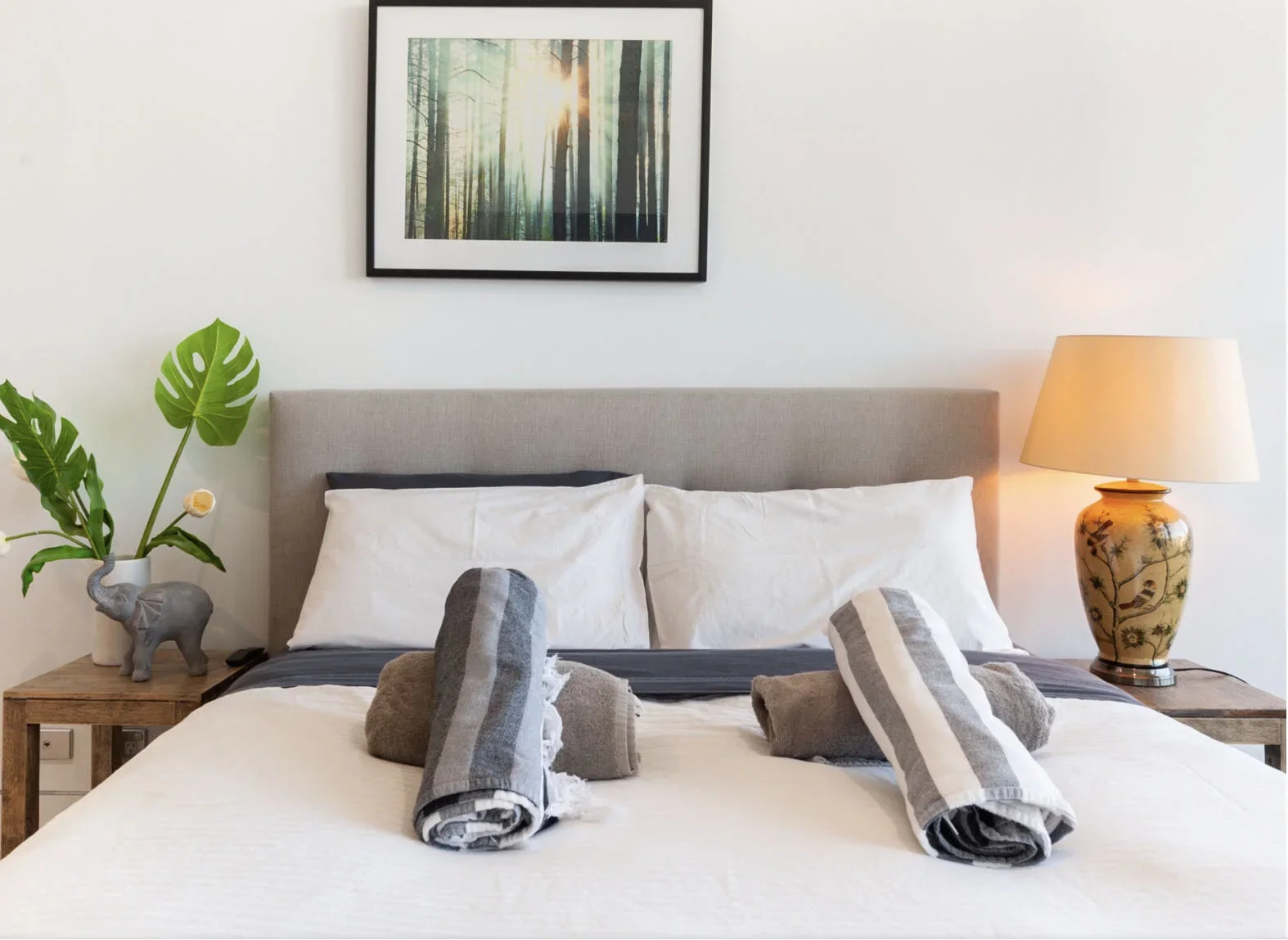 A neatly made bed with white pillows and a beige headboard in a bedroom. Two rolled towels and a gray cloth are placed on the bed. On the left side, a wooden nightstand holds a potted monstera plant and an elephant-shaped figurine. On the right side,