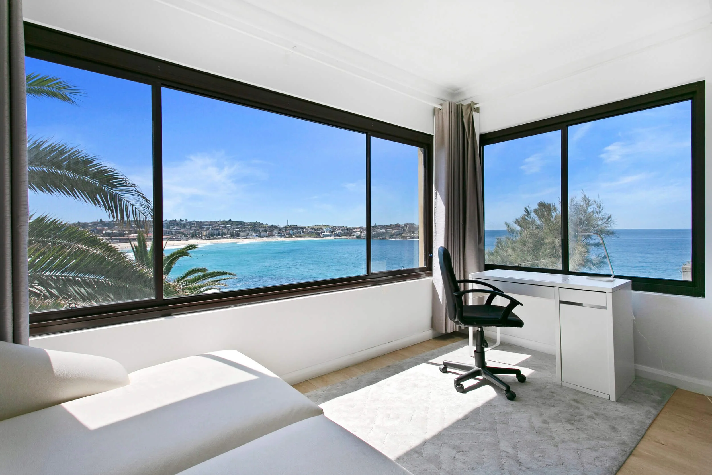 Room with large windows overlooking the ocean, with a white sofa, a white desk, and a black office chair.