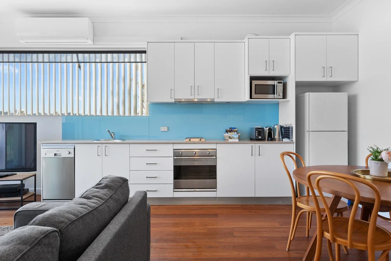 Modern kitchen with white cabinets, stainless steel oven, microwave, toaster, kettle, and fridge, alongside a wooden dining table and chairs, gray sofa, and a window with vertical blinds.