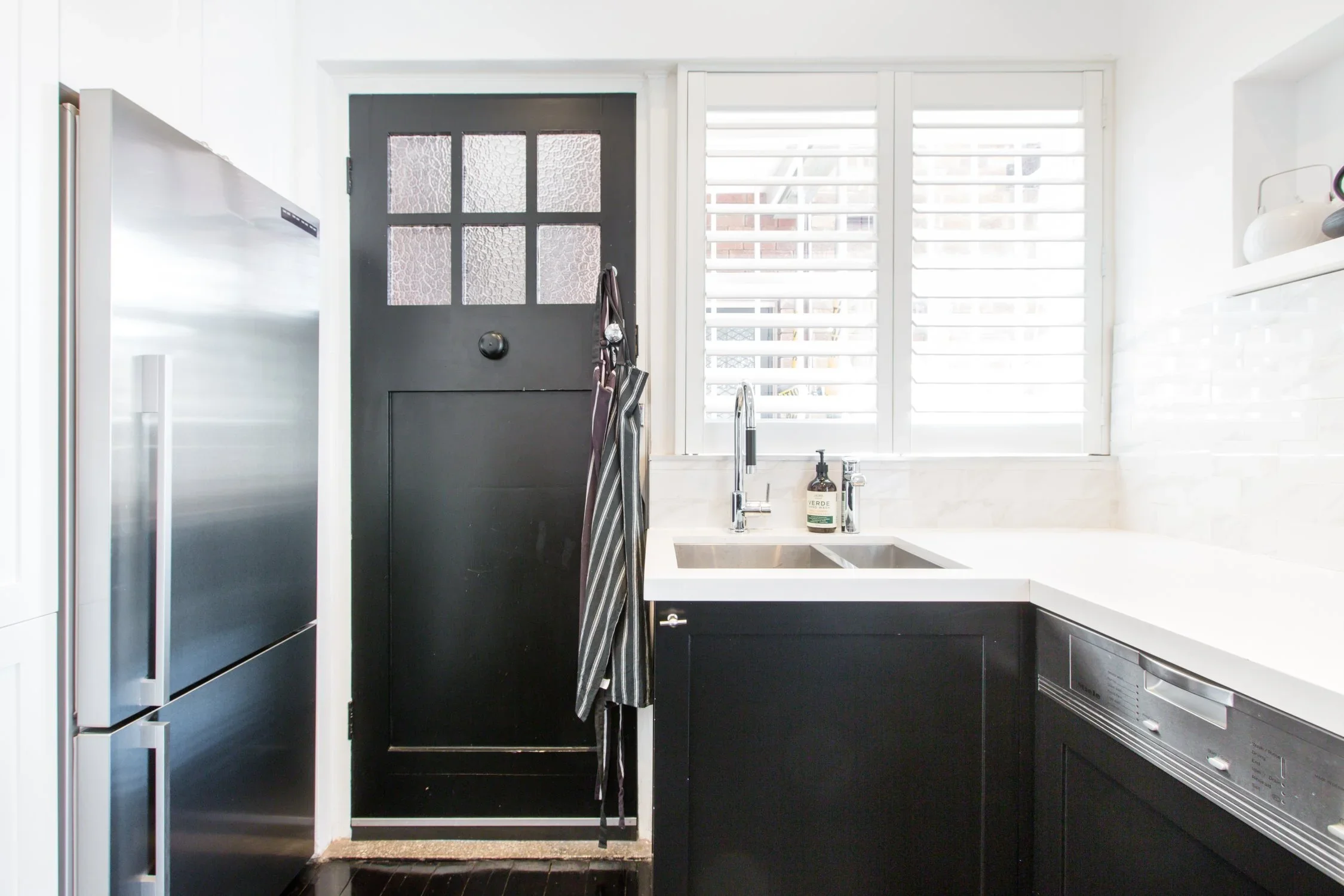 A modern kitchen with a black door, stainless steel refrigerator, black and white cabinetry, white countertops, a window with white blinds, and kitchen accessories.