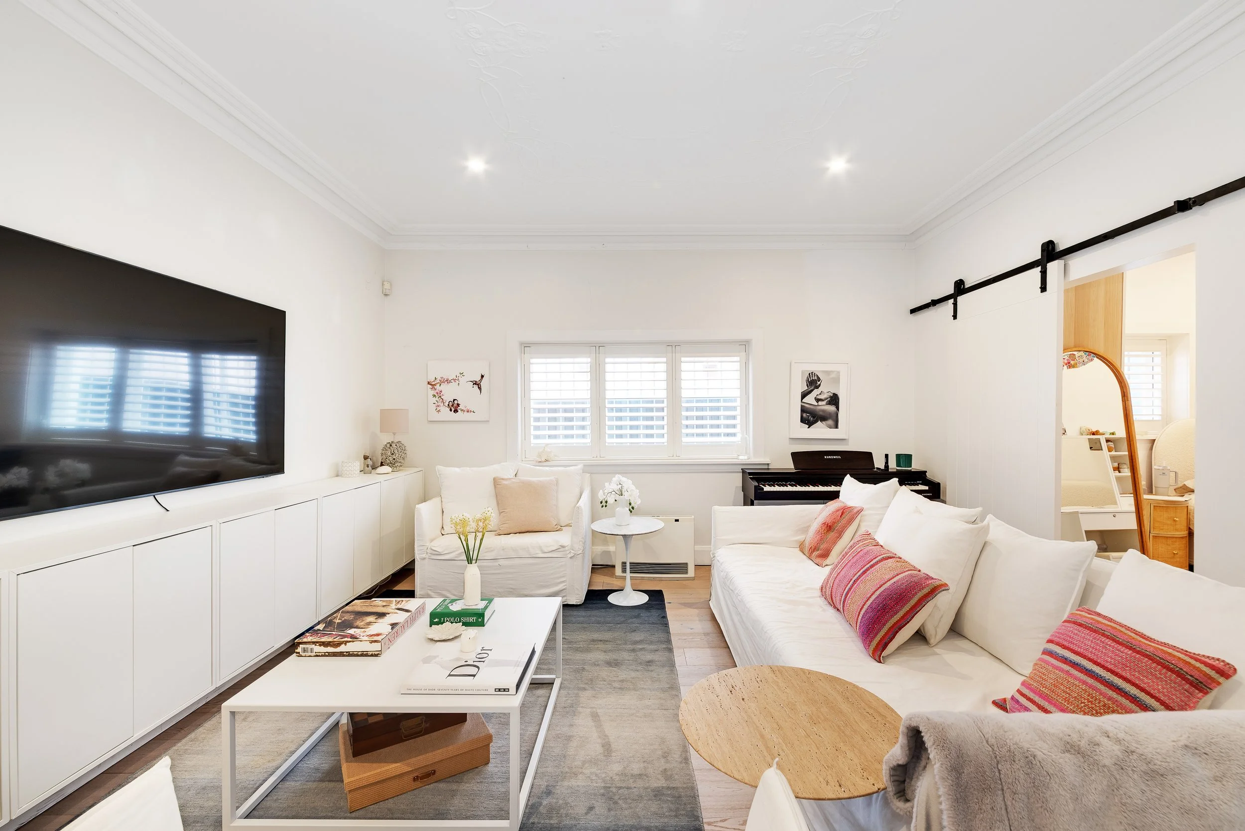 Bright living room with white sofas, a wall-mounted flat-screen TV, a low coffee table with books, large windows with blinds, a light-colored rug, and decorative items.