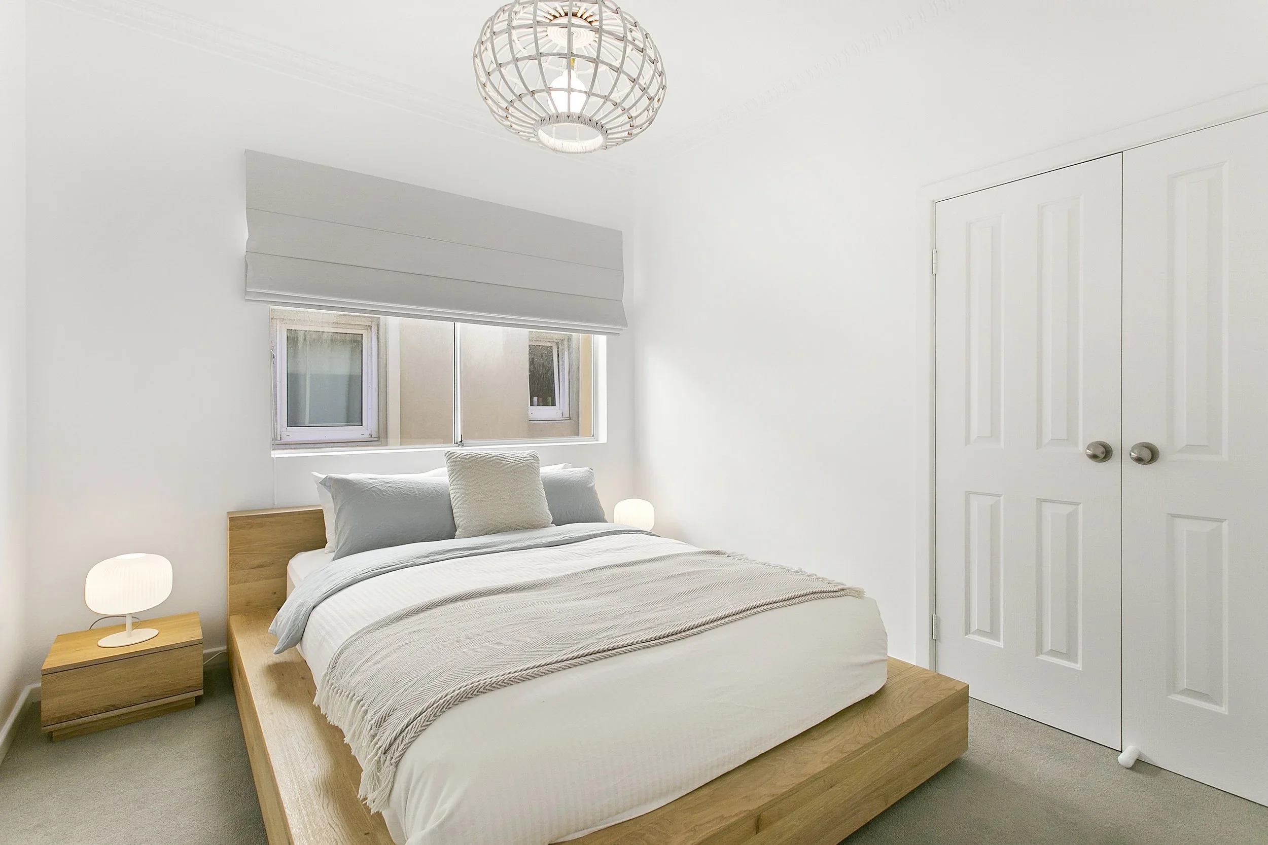 A minimalist bedroom with a wooden bed frame, white bedding, and a beige throw blanket, two bedside lamps, a window with a gray shade, and white closet doors.