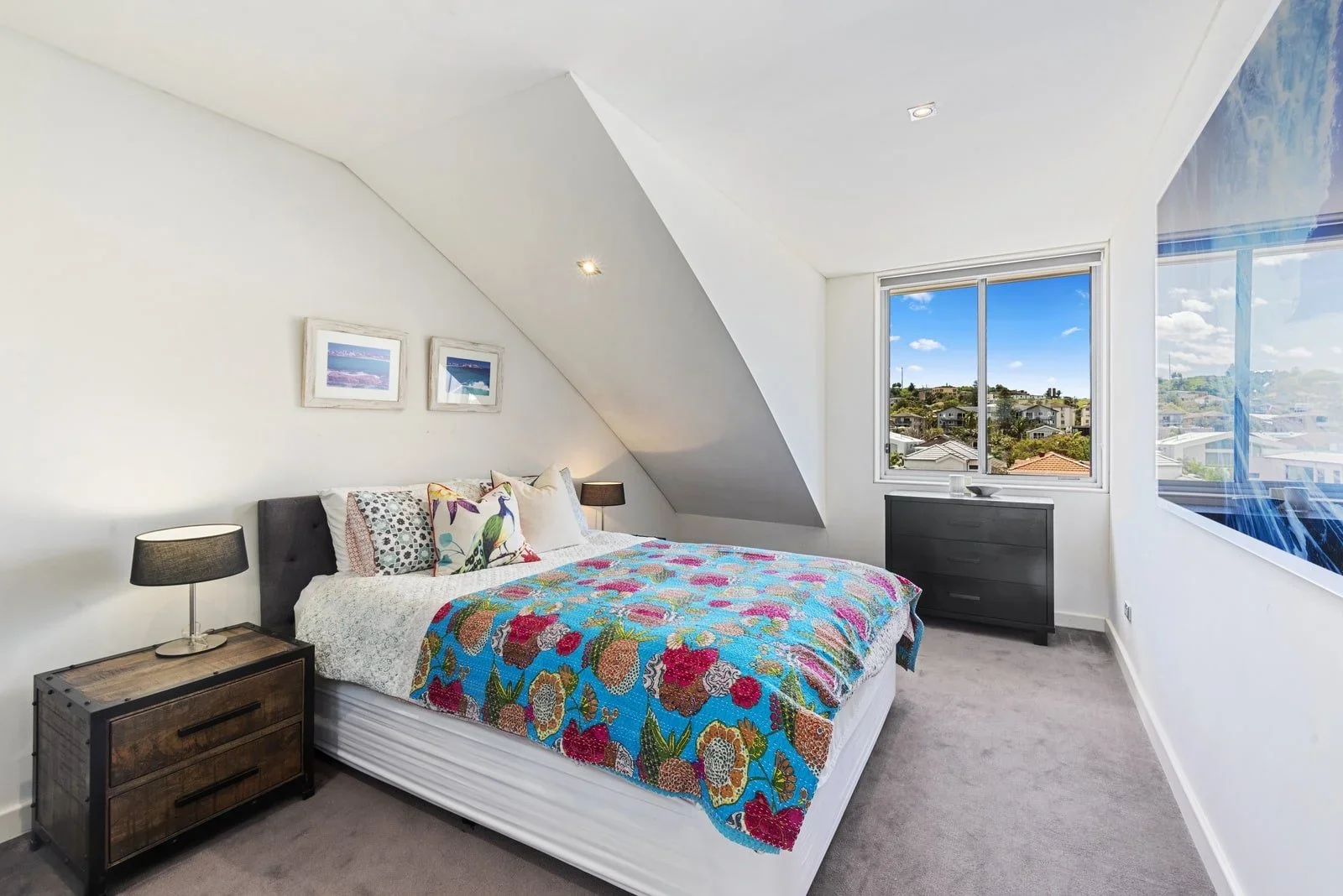 Bright bedroom with sloped ceiling, decorated with a colorful quilt on the bed, framed pictures on the wall, two black bedside lamps, a dark dresser, and a large window showing a view of houses and blue sky.