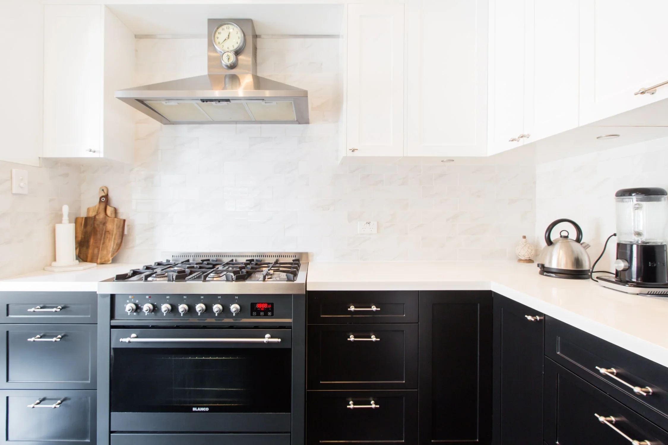 Modern kitchen with black cabinets, white countertops, stainless steel stove and range hood, wall clock, and kitchen appliances on counter.