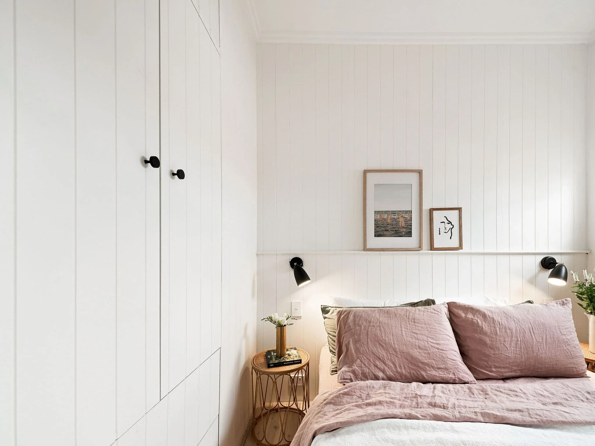 A cozy bedroom with a white paneled wall, framed pictures, black wall-mounted reading lamps, pink bed linens, a bedside table with a plant, and a built-in closet with black knobs.