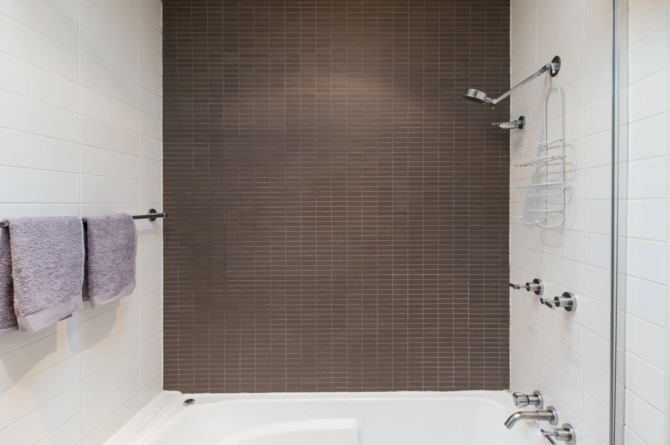 Modern shower with brown tiled wall, white tiled side walls, two purple towels on a towel bar, and shower fixtures including a showerhead, handles, and a wire shower caddy.