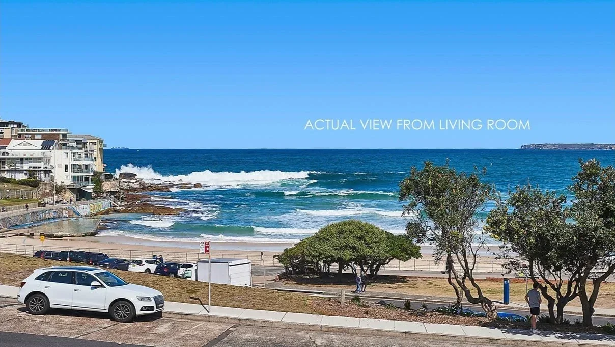 View of a beach with blue ocean waves, a sandy shore, and partly cloudy sky, with parked cars and trees in the foreground along a coastal road.