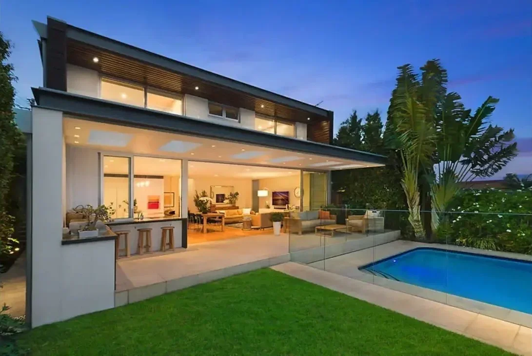 Modern house with large glass windows, interior living room visible, outdoor patio, swimming pool, lush green lawn, trees, and evening sky.