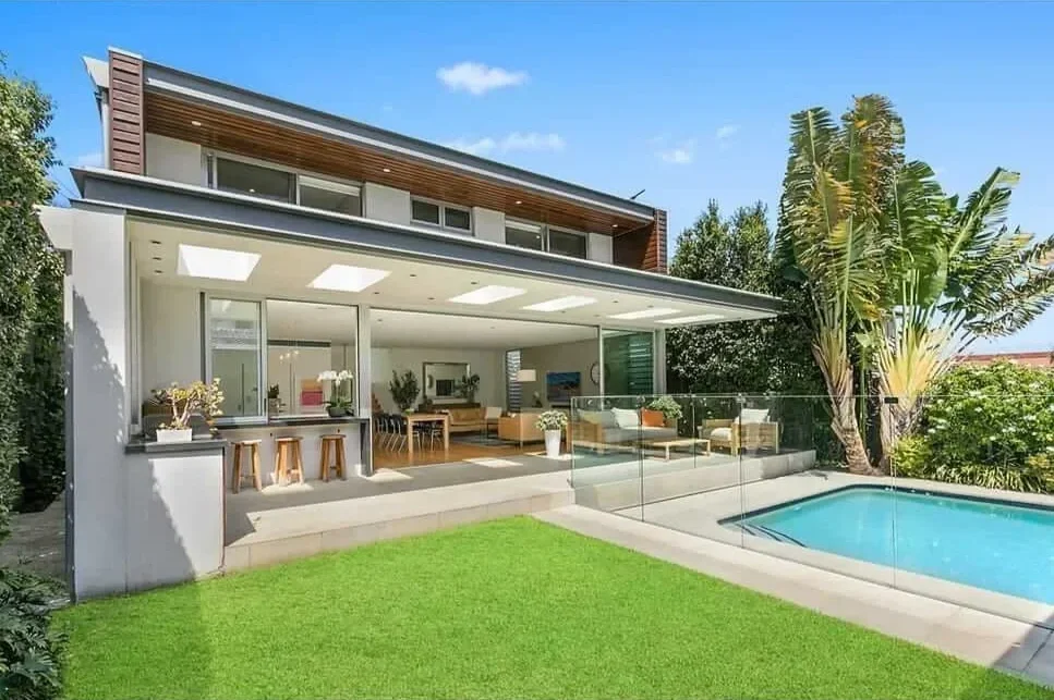 Modern two-story house with an outdoor patio, glass railing, swimming pool, and lush greenery under a blue sky.