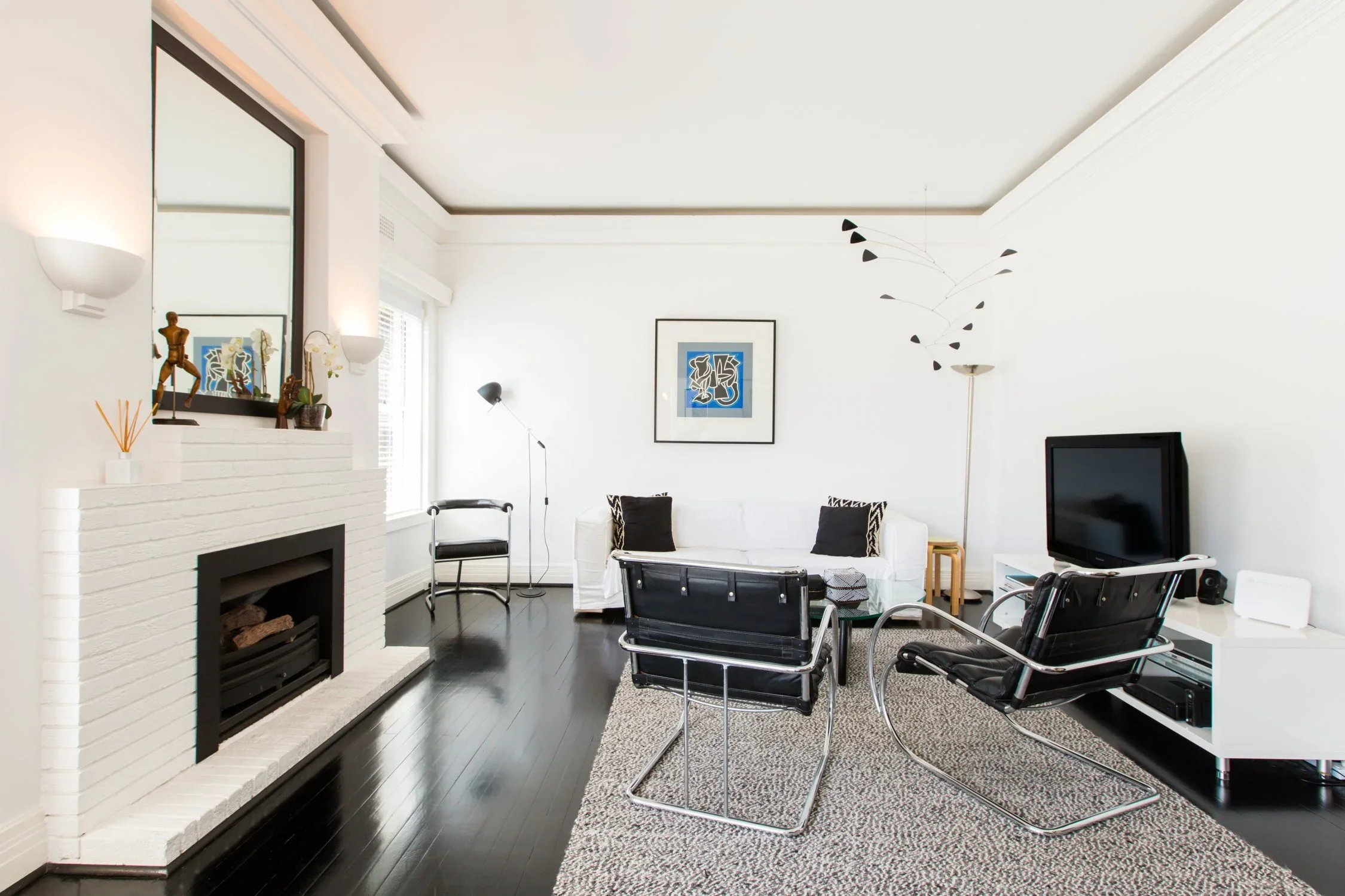 Living room with white walls, black hardwood floors, a white fireplace, a white sofa, two black leather chairs, and a TV on a white media console. Decorations include framed artwork, a floor lamp, and small plants.