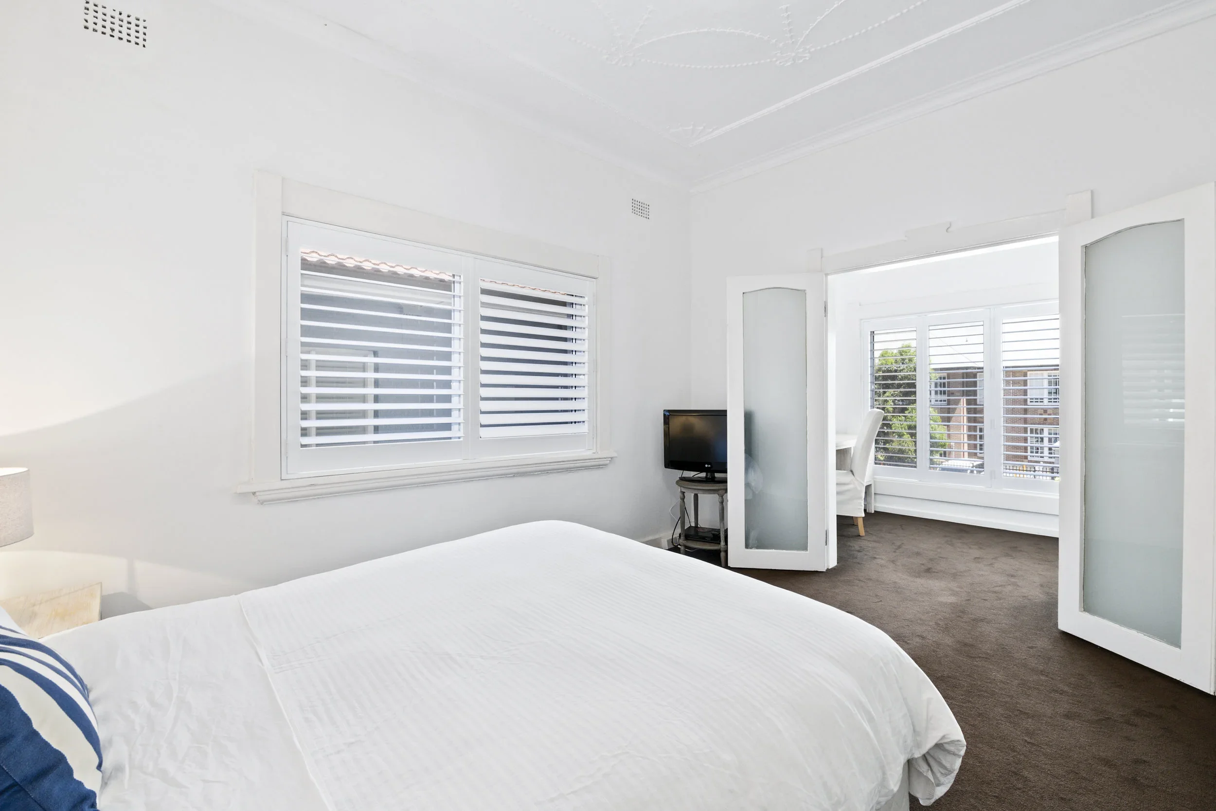 Bright white bedroom with a bed, windows with blinds, and an adjoining sitting area with a chair and a small television