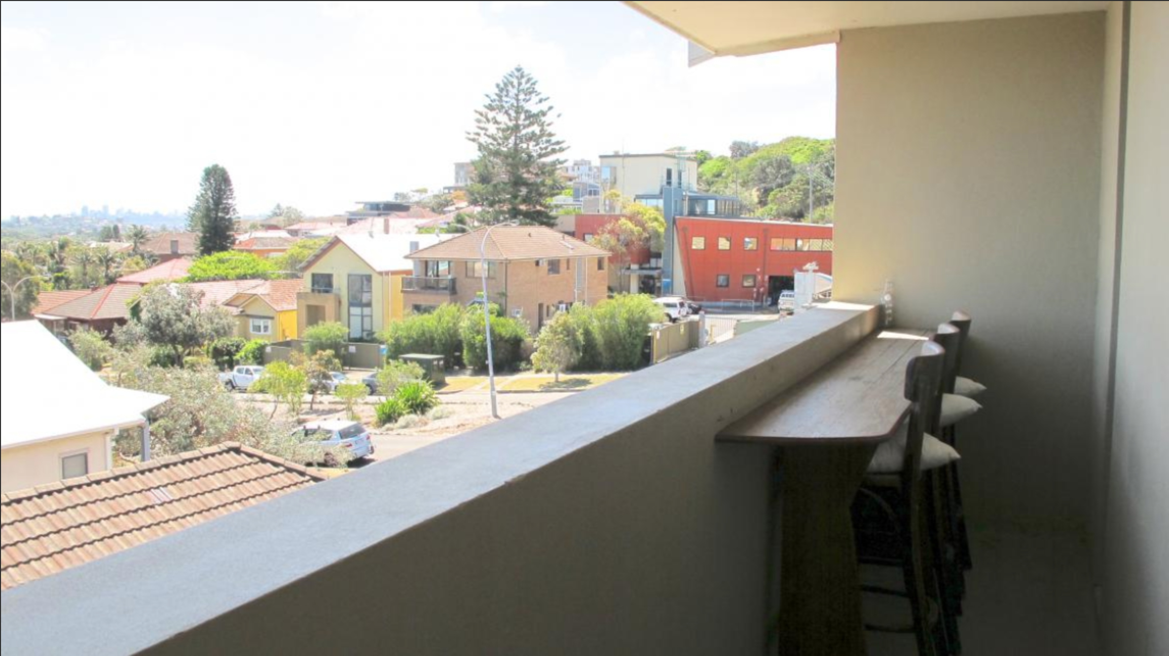 View from a balcony showing residential neighborhood with colorful houses, trees, parked cars, and a streetlamp under a clear sky.
