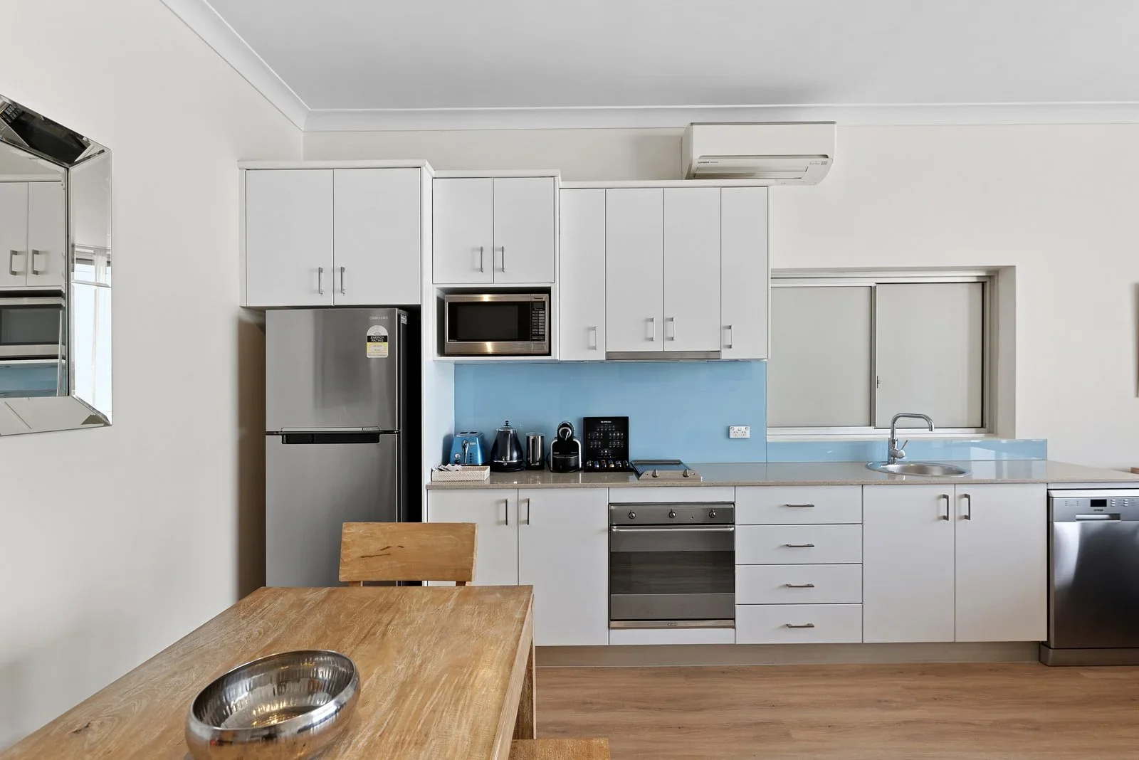 Modern kitchen with white cabinets, stainless steel appliances, wooden dining table, and a light blue backsplash.