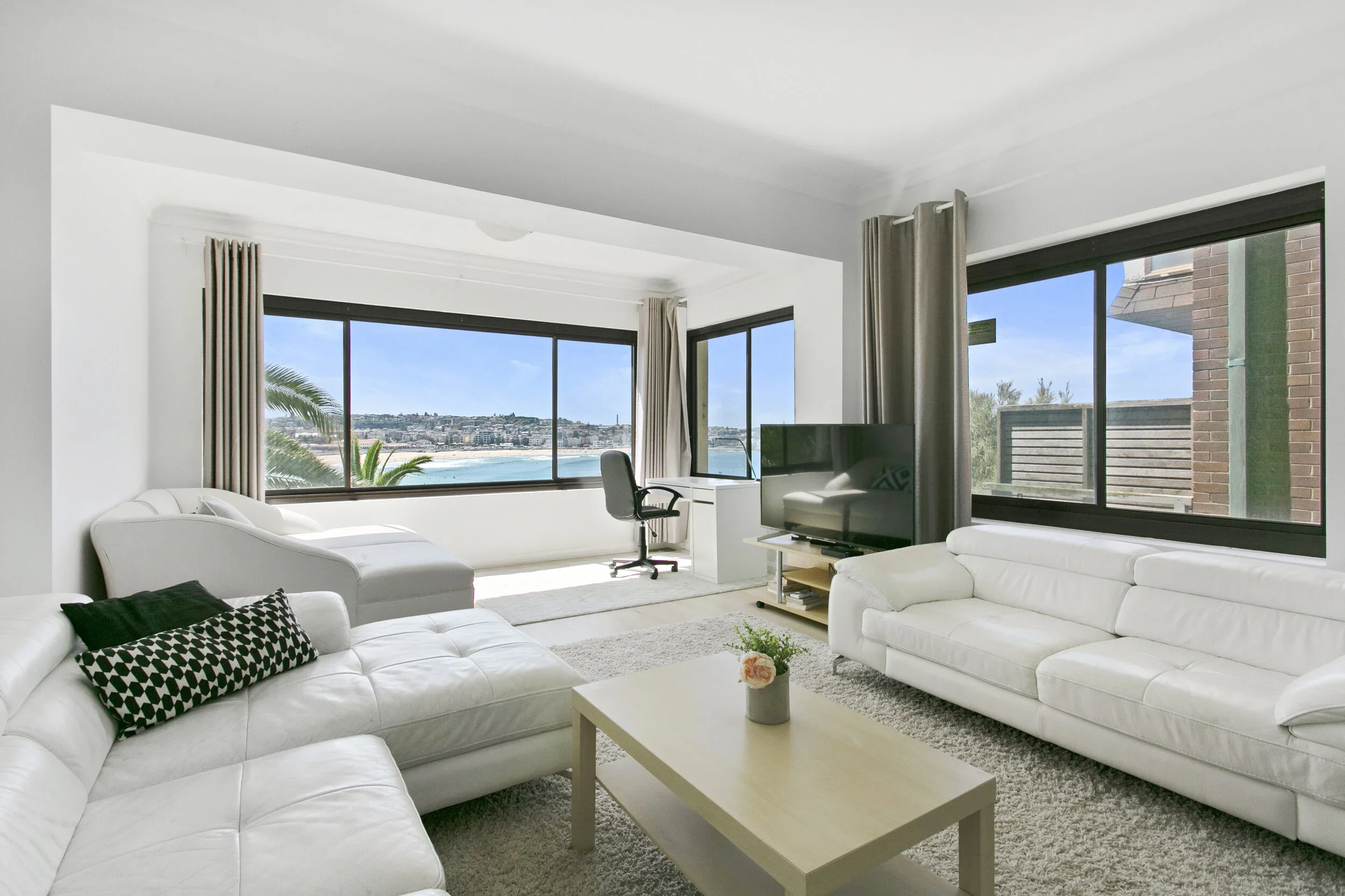 Living room with white leather sectional sofa, flat-screen TV, and large windows showing beach and ocean view.