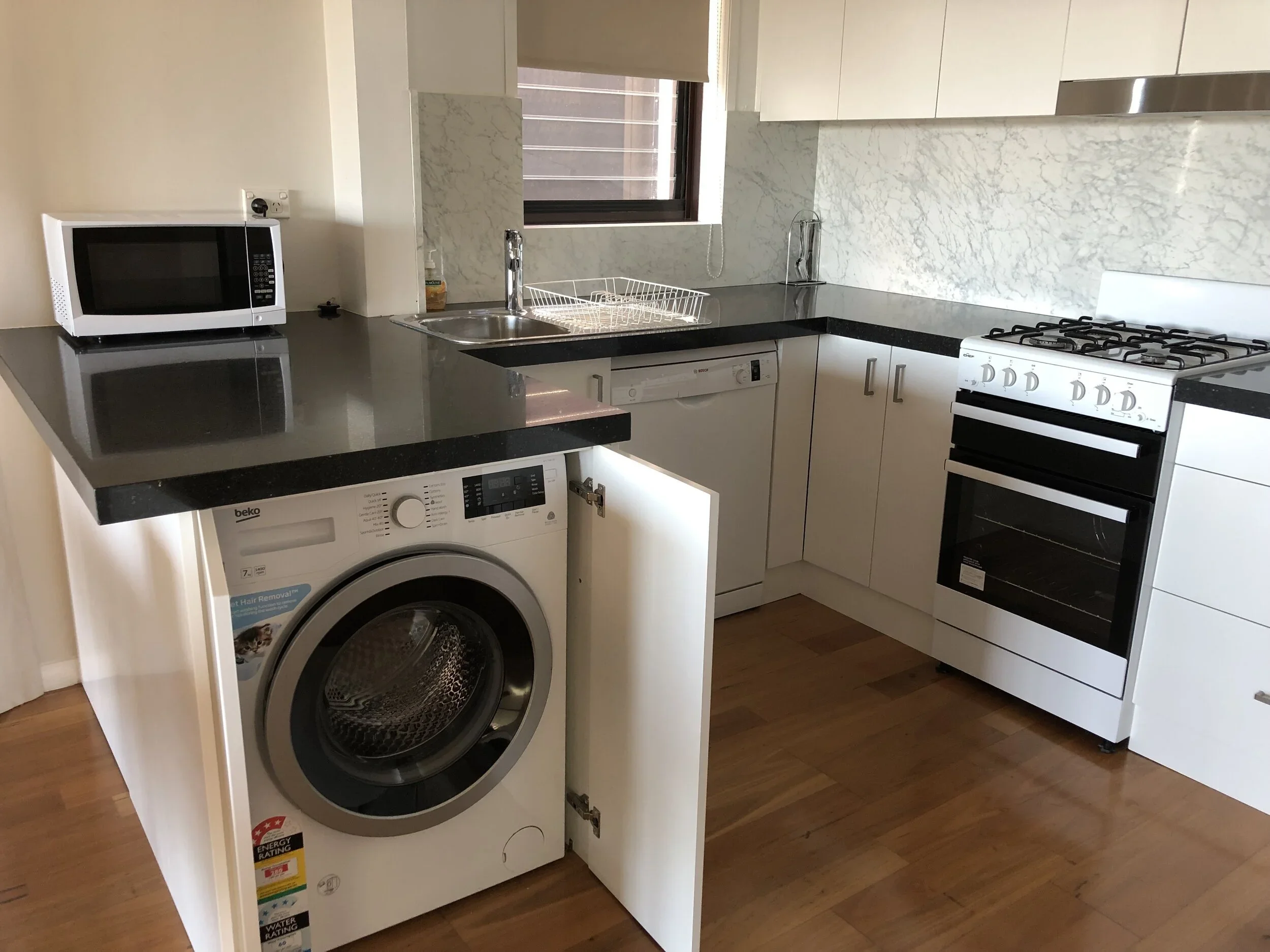 Kitchen with a microwave on the counter, washing machine in an open cabinet, sink with faucet, dishwasher, and a stove along white cabinets and marble backsplash, with hardwood floor.
