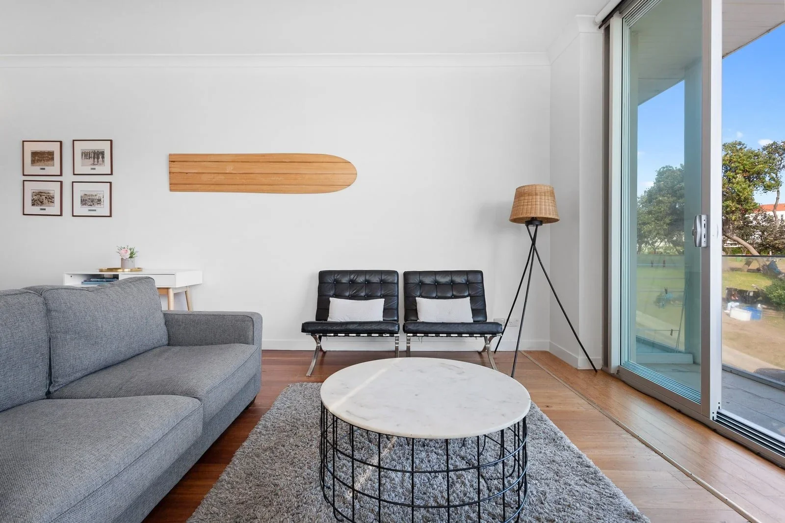 Living room with gray sofa, black chairs, white wall, wooden wall art, floor lamp, sliding glass door, and view of greenery outside.