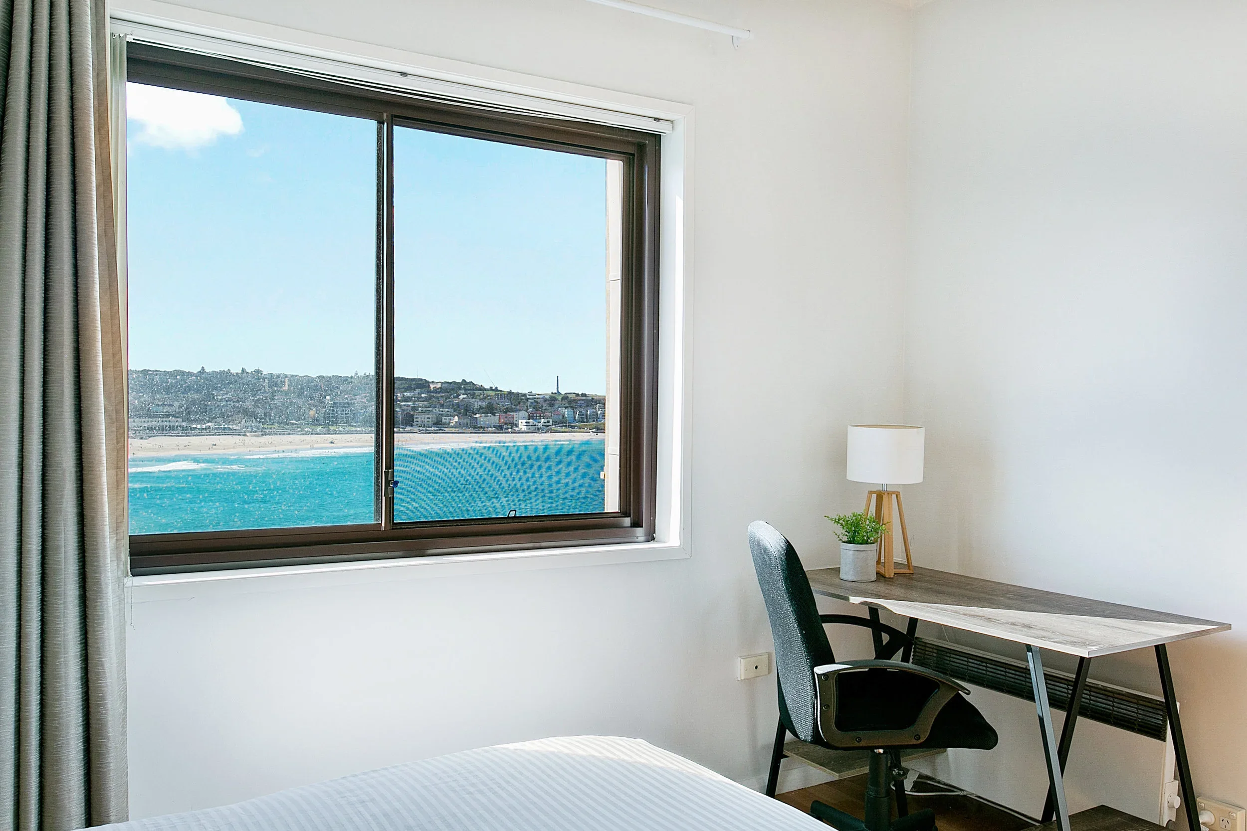Room with a window overlooking the beach and ocean, a desk with a lamp and potted plant, and an office chair.