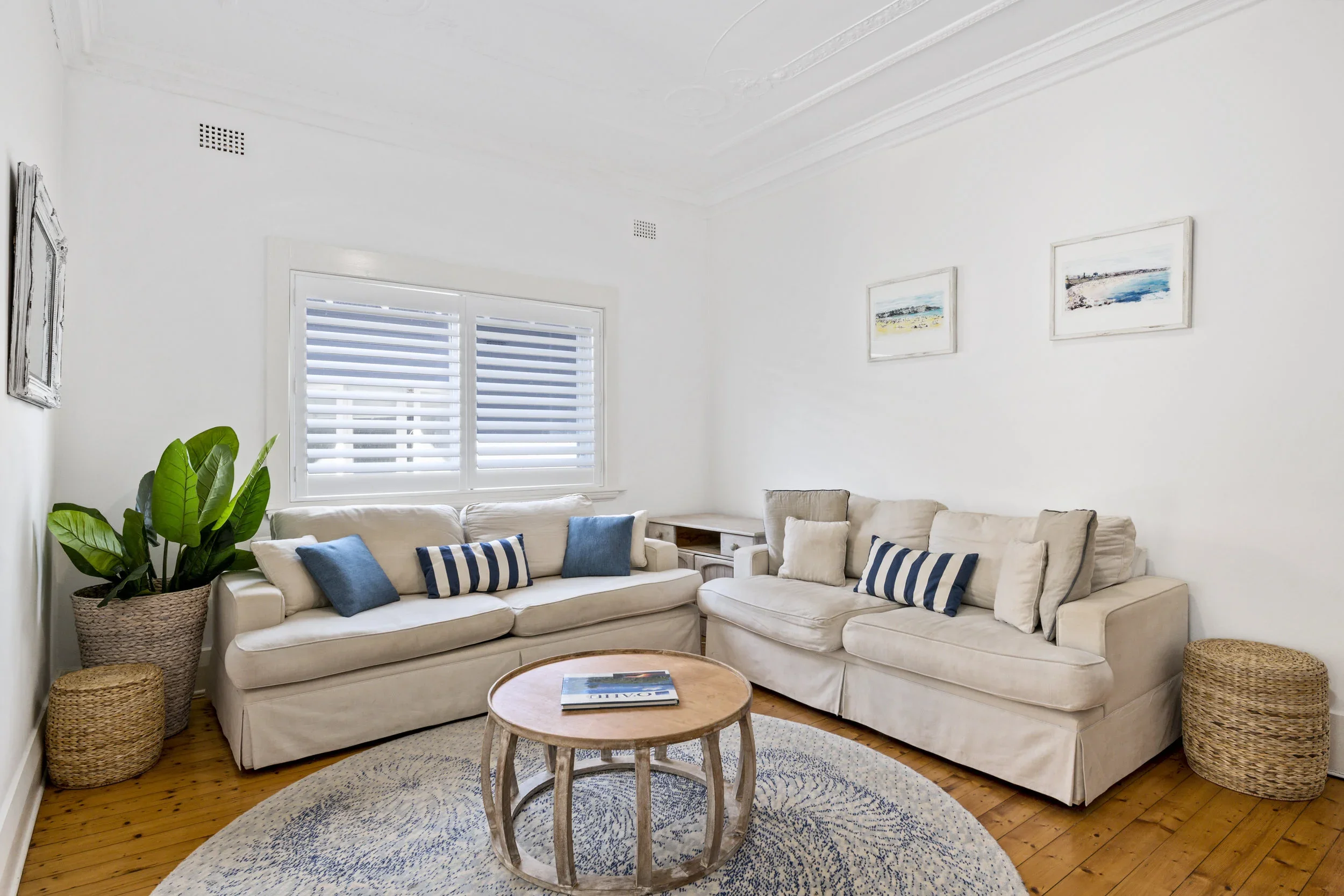 Living room with white sofas, blue and beige pillows, a round wooden coffee table, a potted plant, and framed wall art in a bright space.