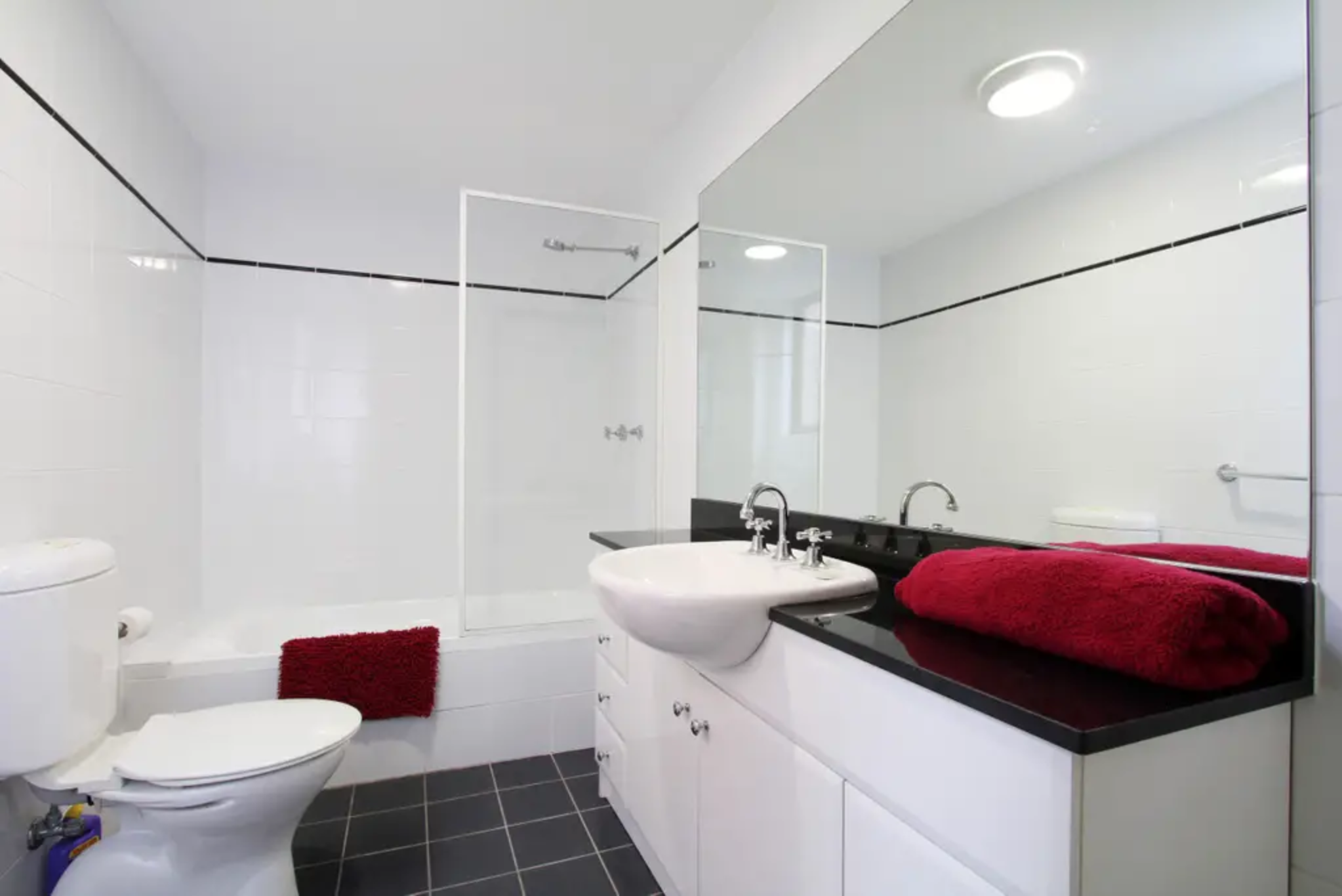 Bathroom with white tiled walls, black trim, a toilet, a red towel on the floor, a sink with a black countertop, a large mirror, and a glass-enclosed shower.