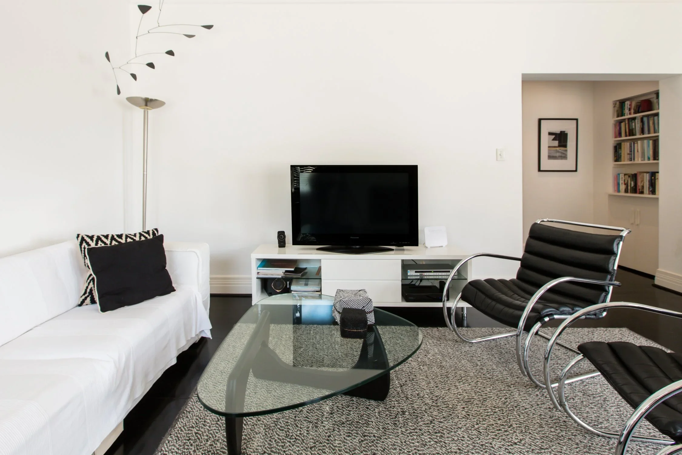Living room with white sofa, black accent pillows, glass coffee table, black lounge chairs, and a white TV console with a flat-screen TV, bookshelves nearby, and a patterned rug.