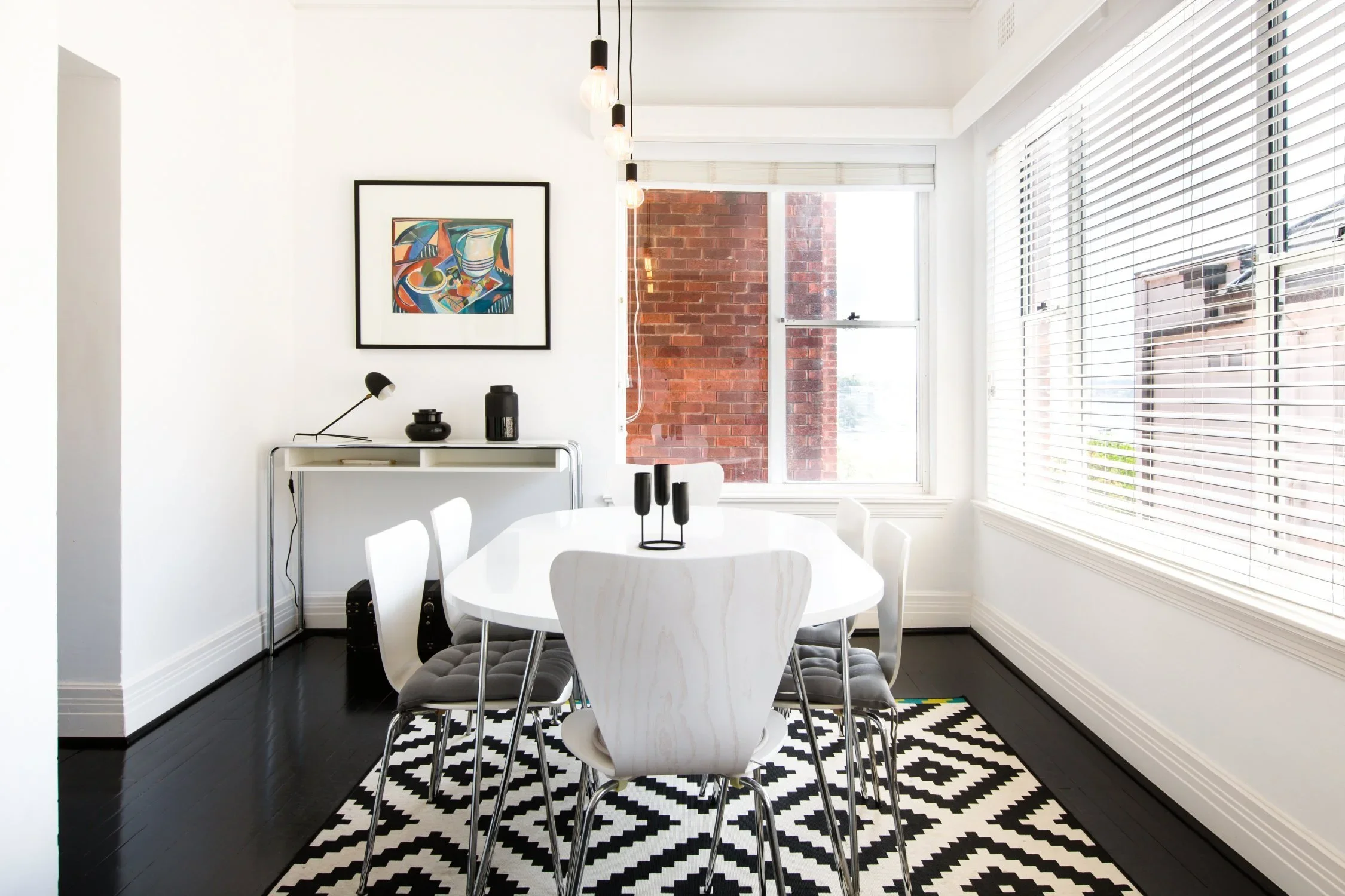Bright dining room with white walls, black and white geometric patterned rug, white dining table with six chairs, large windows with blinds, colorful abstract artwork on the wall, and modern decor including a black vase and candlesticks.