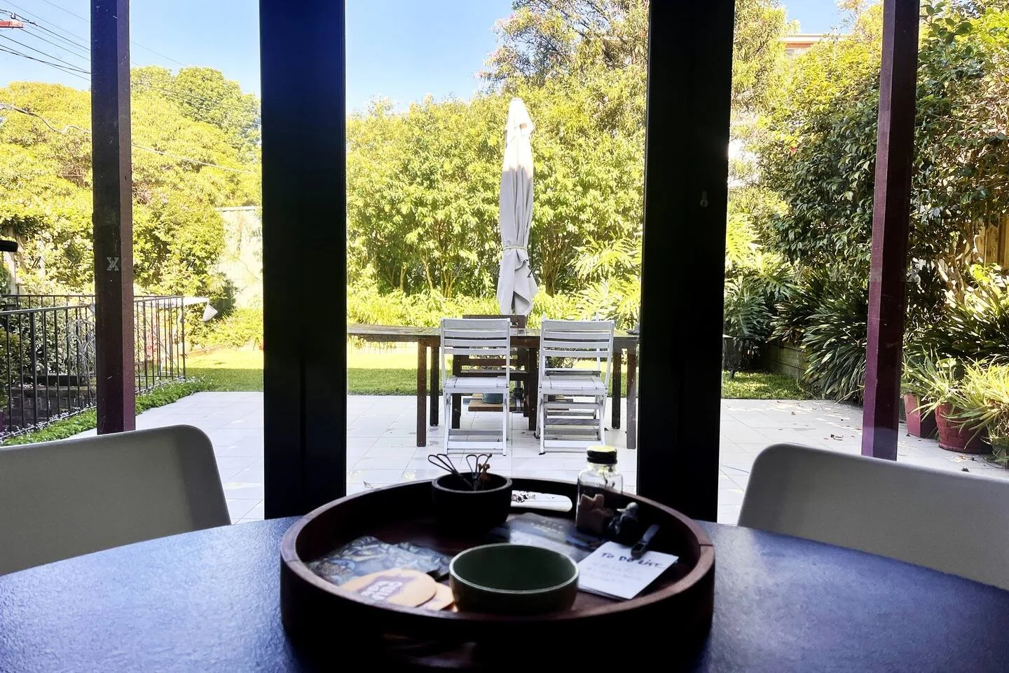 View of a backyard patio through a glass sliding door, with outdoor table, chairs, and a white umbrella, surrounded by lush greenery.