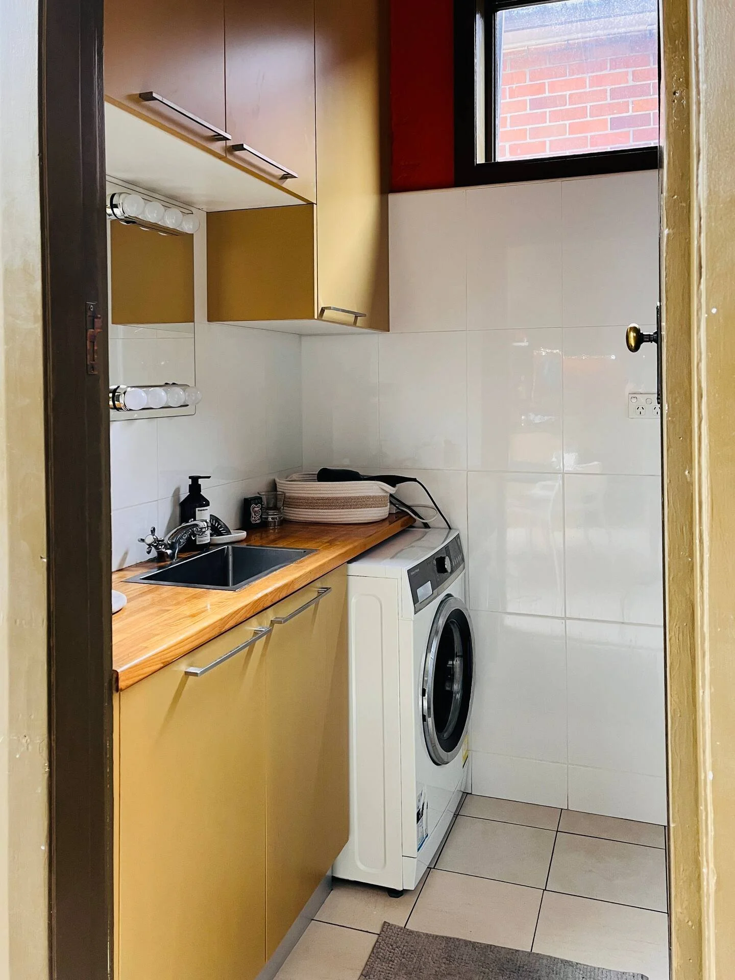 A small laundry room with a washer, a wooden countertop, yellow cabinets, a small sink, and a window with brick wall visible outside.