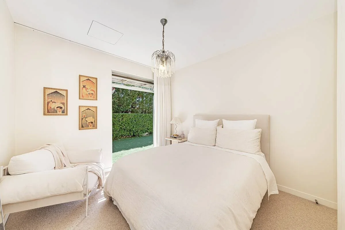 A bedroom with white walls, a white bed with white pillows, a small white nightstand with a lamp, a white sofa with a throw blanket, and framed artwork on the wall. Sliding glass door leads outside to a green hedge and lawn.