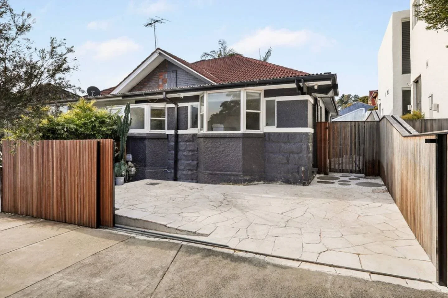 Backyard of a house with a concrete patio, wooden fences on both sides, a dark-colored house with white-framed windows, and a neighboring white modern building partially visible.