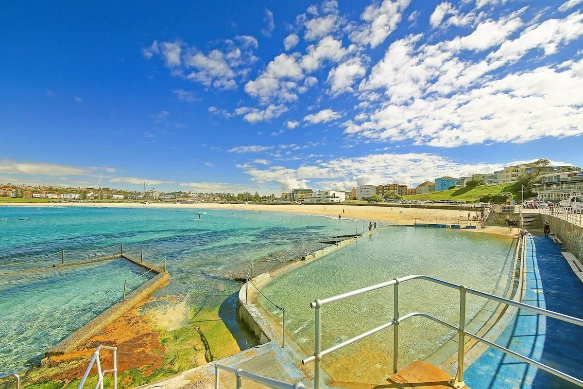 Beach with a clear swimming pool in the foreground, sandy shoreline with a few people, and colorful residential buildings under a partly cloudy blue sky.