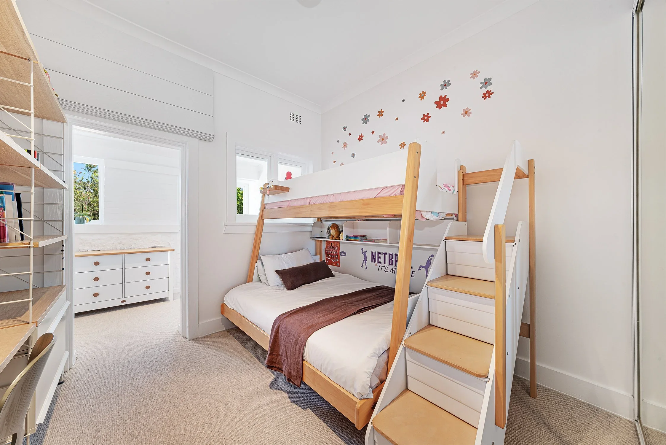 Bedroom with a bunk bed and stairs, decorated with flower stickers on the wall, with windows and bookshelf nearby.