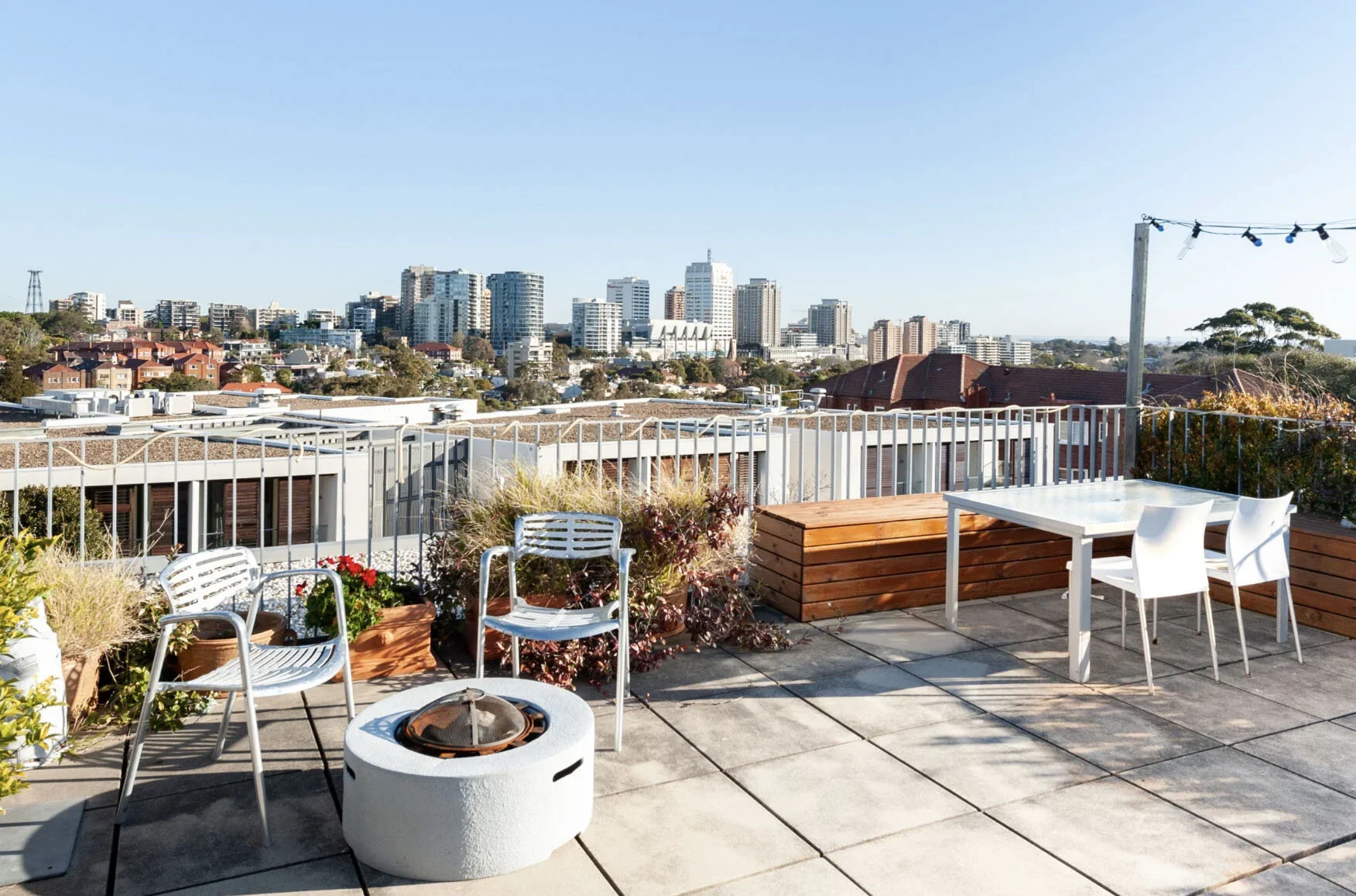 Rooftop patio with outdoor furniture, chairs, table, and plants, overlooking a city skyline with tall buildings and clear blue sky.
